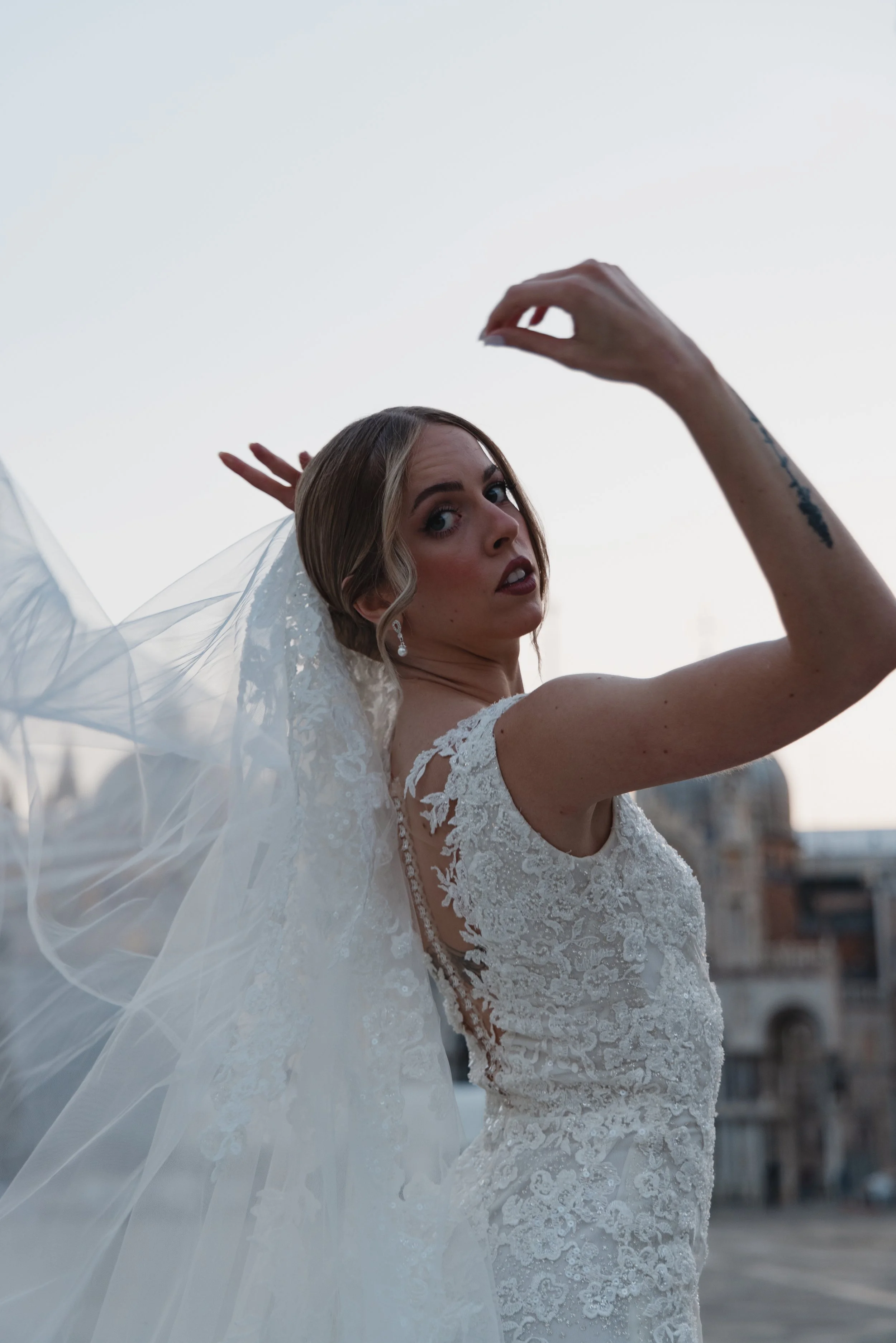 A woman in a white lace wedding dress with a veil, posing outdoors, looking over her shoulder with her arm raised, against a city skyline background during sunset.
