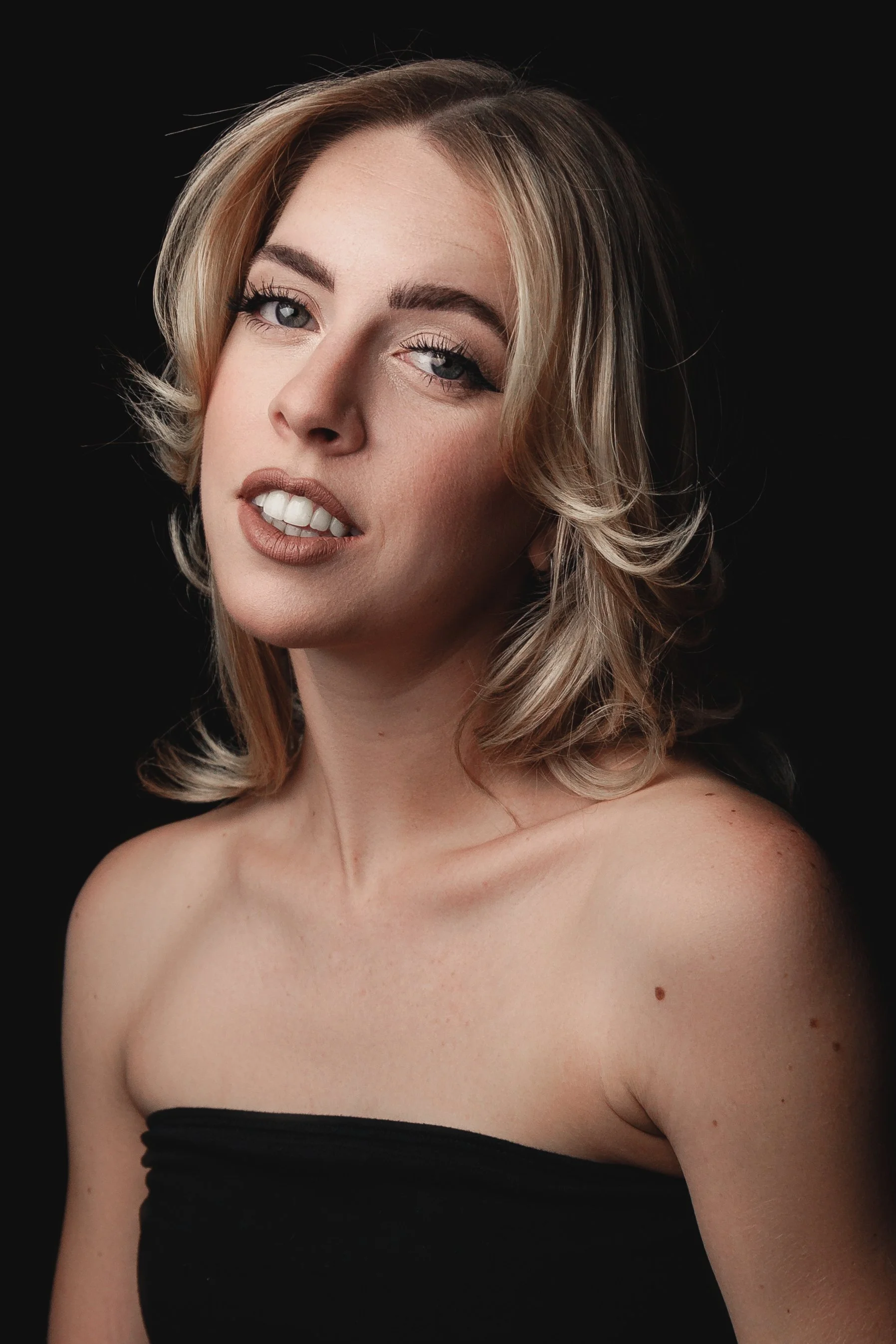 A woman with shoulder-length blonde hair styled in loose waves, wearing makeup with defined eyebrows, eyeshadow, mascara, and nude lipstick, posing against a black background. She has no shirt or dress, only a black strapless garment, revealing her s