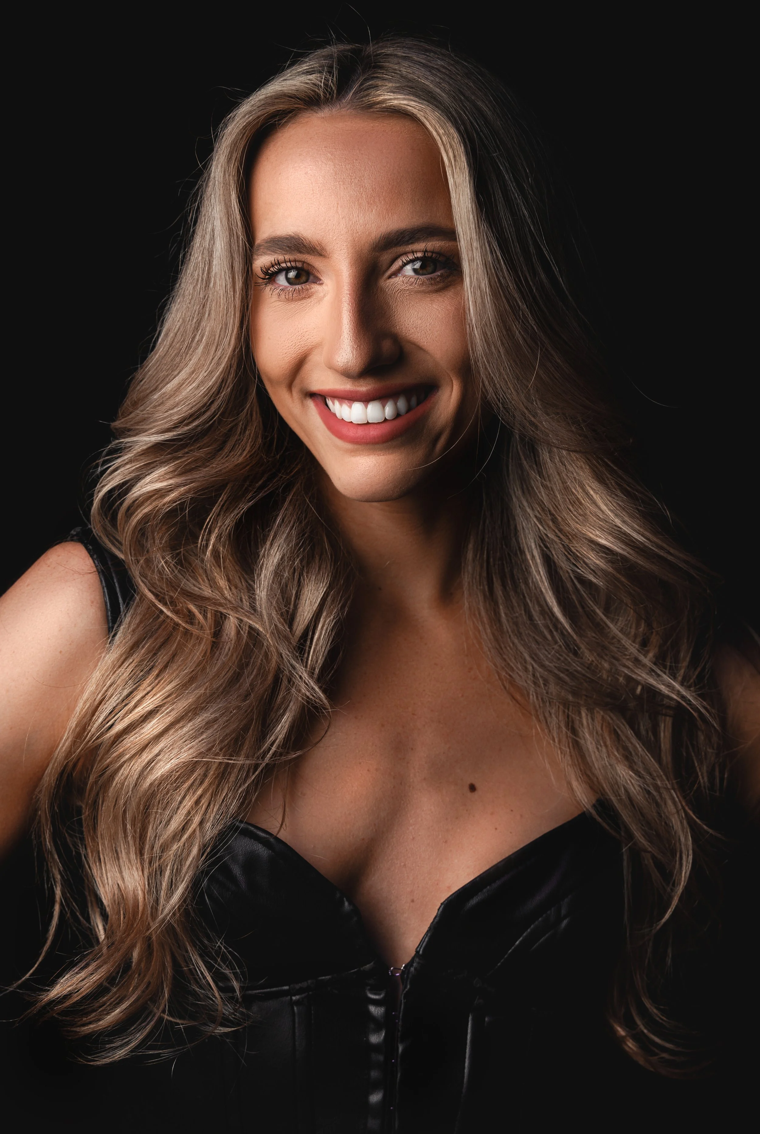 Close-up portrait of a smiling woman with long wavy blonde hair, wearing a black leather top against a black background.
