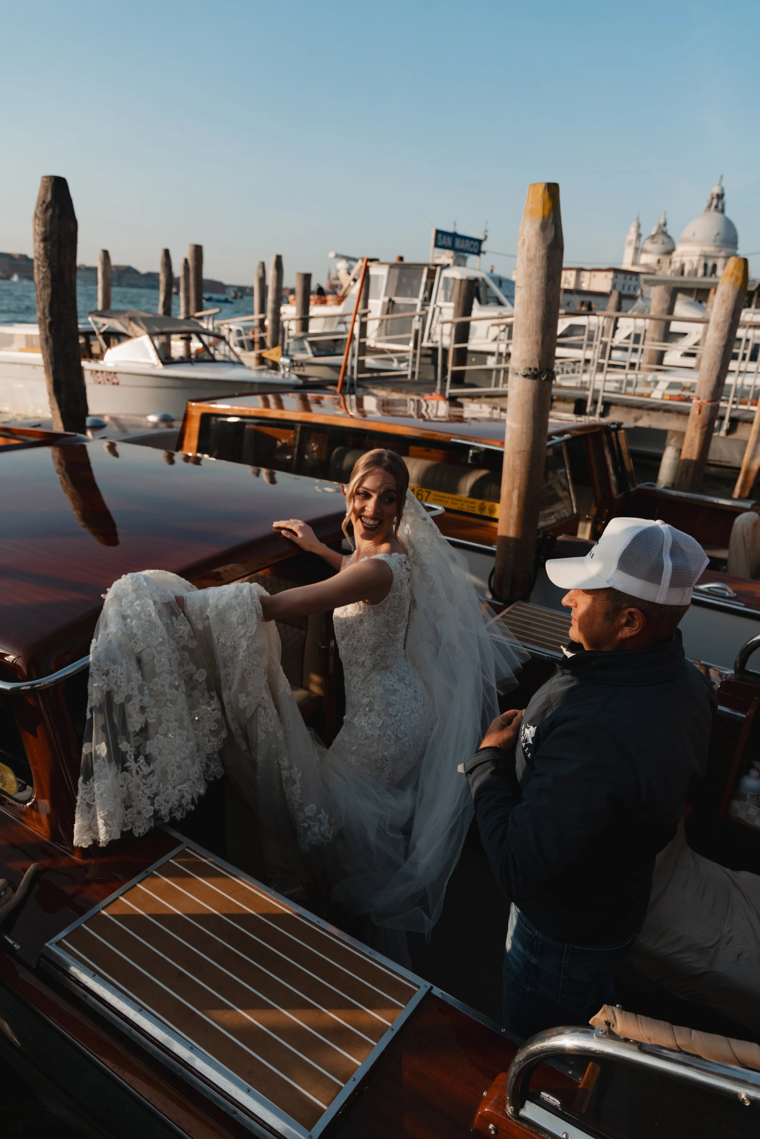 A woman in a wedding dress sitting on a boat with a man, with boats and a cityscape in the background.