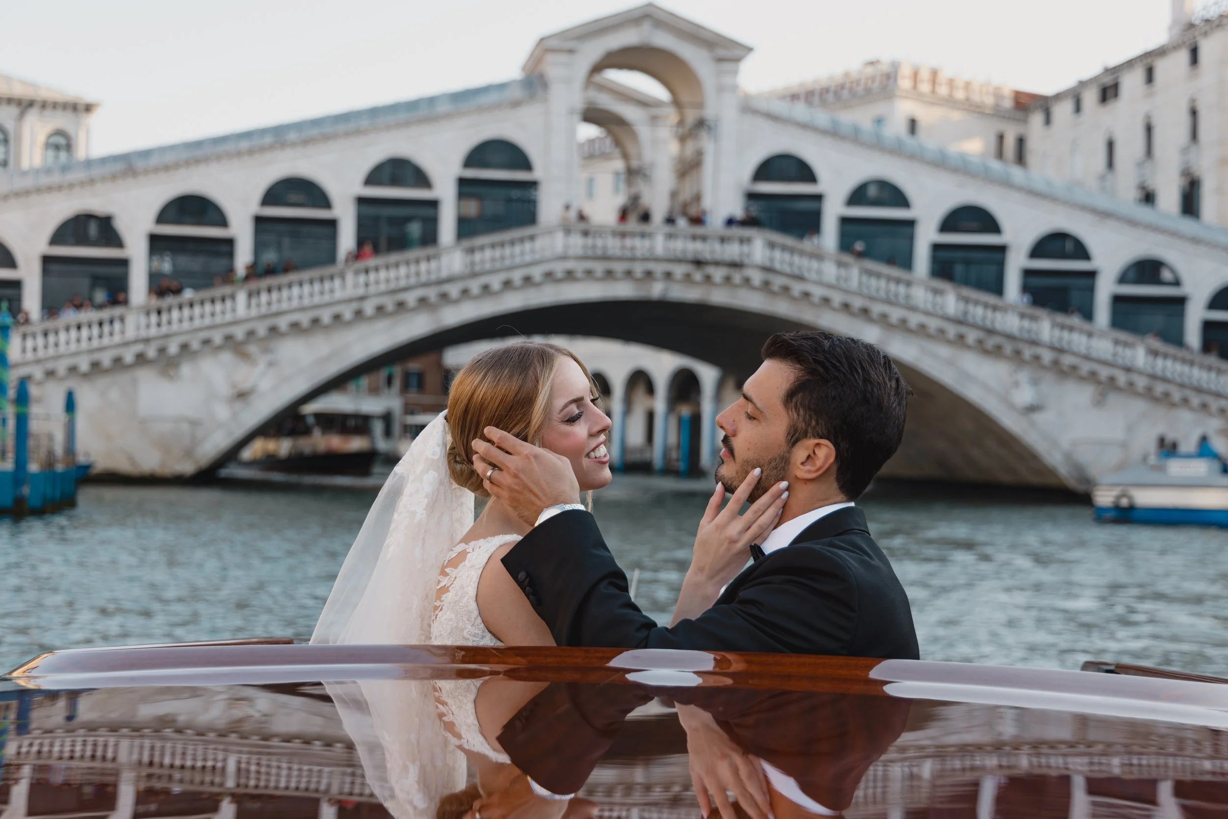 A bride and groom in wedding attire smiling and touching faces while on a boat in Venice, Italy, with the Rialto Bridge in the background.