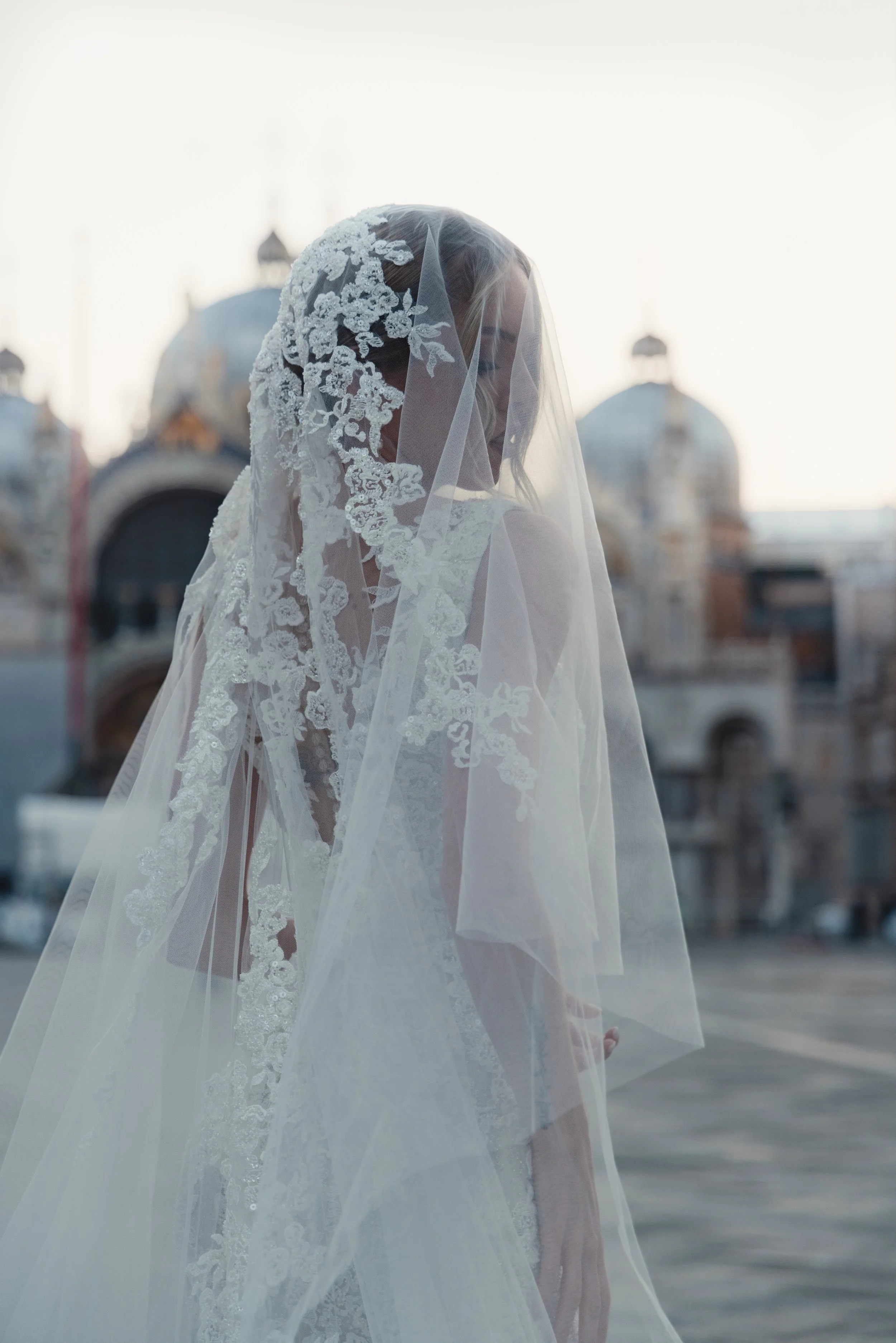 A bride wearing a white lace wedding dress and veil stands outdoors in front of domed architecture, with her head slightly bowed and her face partially obscured by the veil.