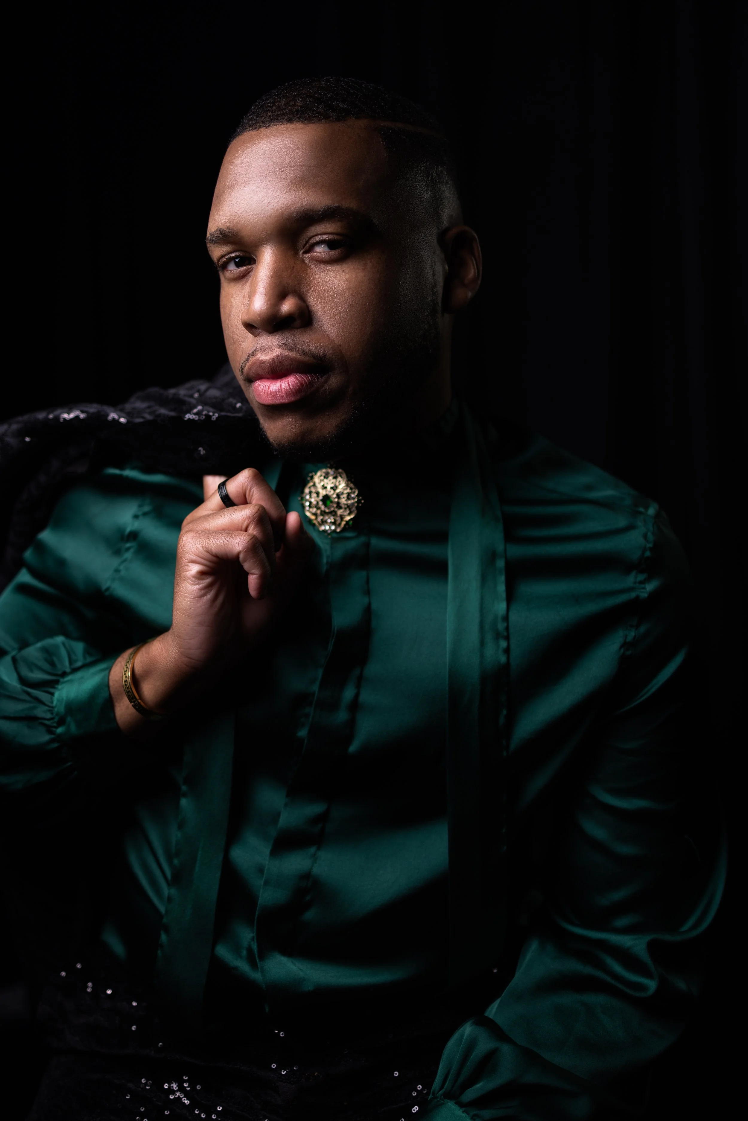 Portrait of a young man with short hair, wearing a teal satin shirt, black sequin pants, and a decorative brooch at the collar, holding a black sequined jacket over his shoulder, against a black background.