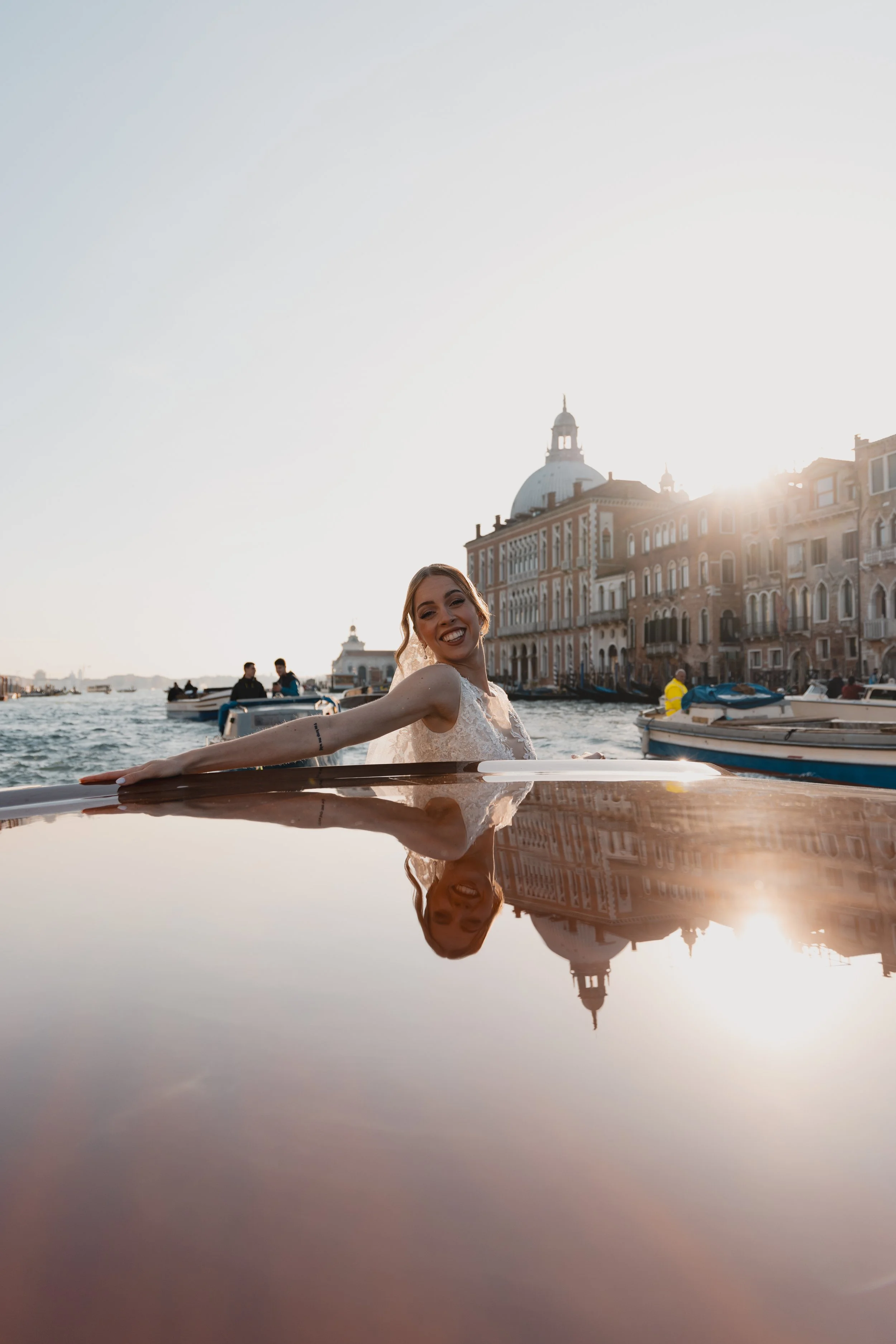 A smiling woman in a white dress on a boat in Venice, Italy, with historic buildings and the dome of a church in the background, during sunset.