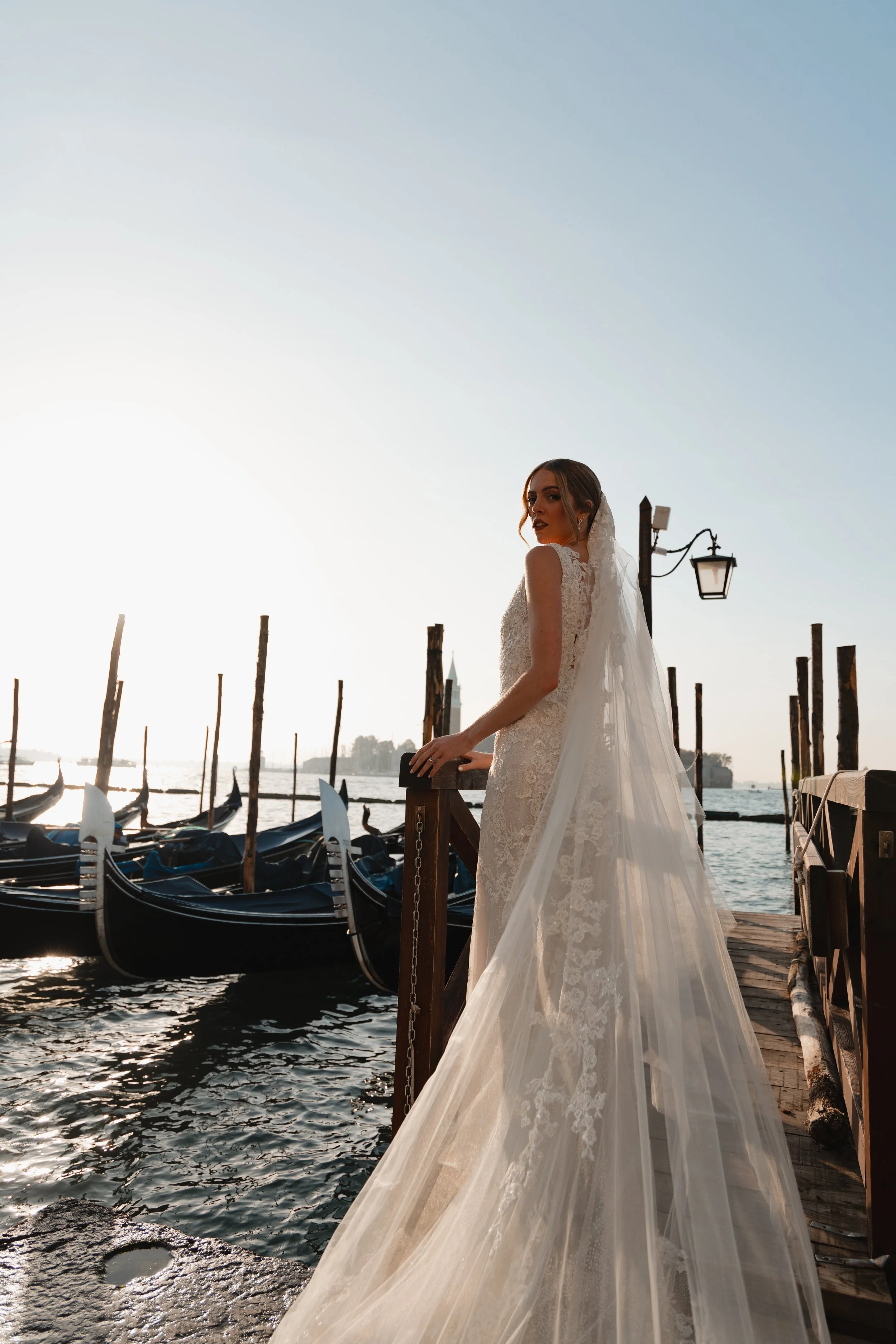 A bride in a lace wedding gown and veil standing on a wooden dock beside boats docked by the water, with a lamp post and a city skyline in the background during sunset.