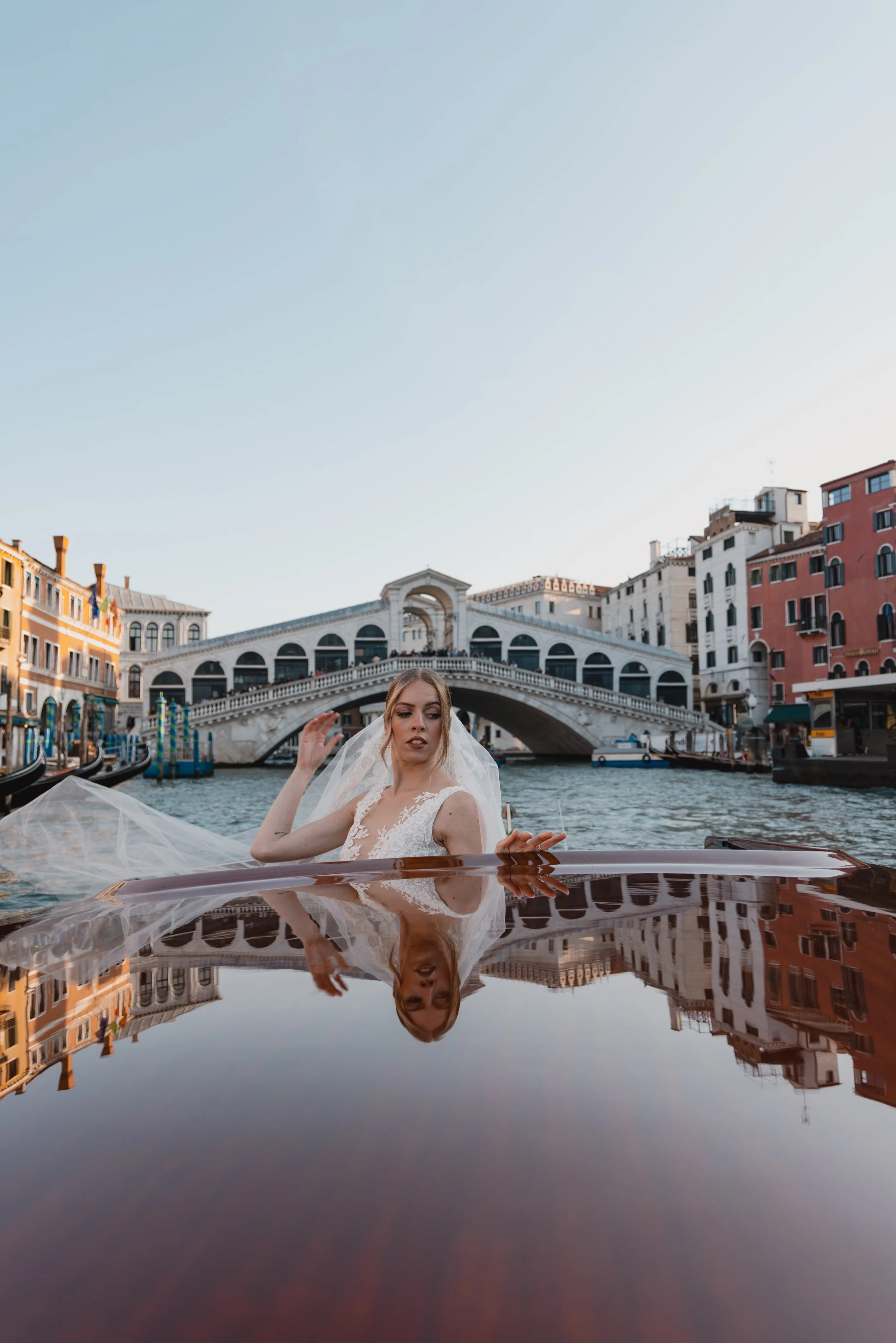A woman in a wedding dress on a boat, with buildings and a bridge in Venice, Italy, in the background, and her reflection visible on the water.