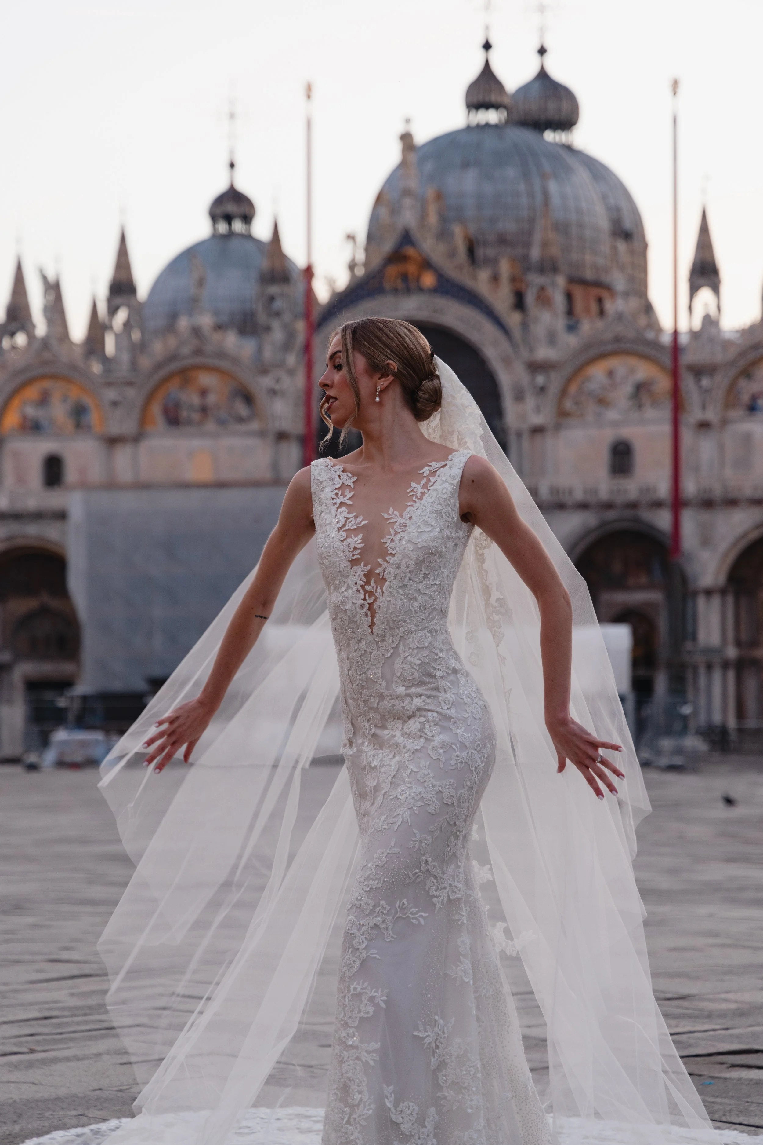 A bride in a white lace wedding dress with a deep V neckline and sheer lace detailing, standing outdoors in front of a historic European-style building with domes and spires, during sunset.