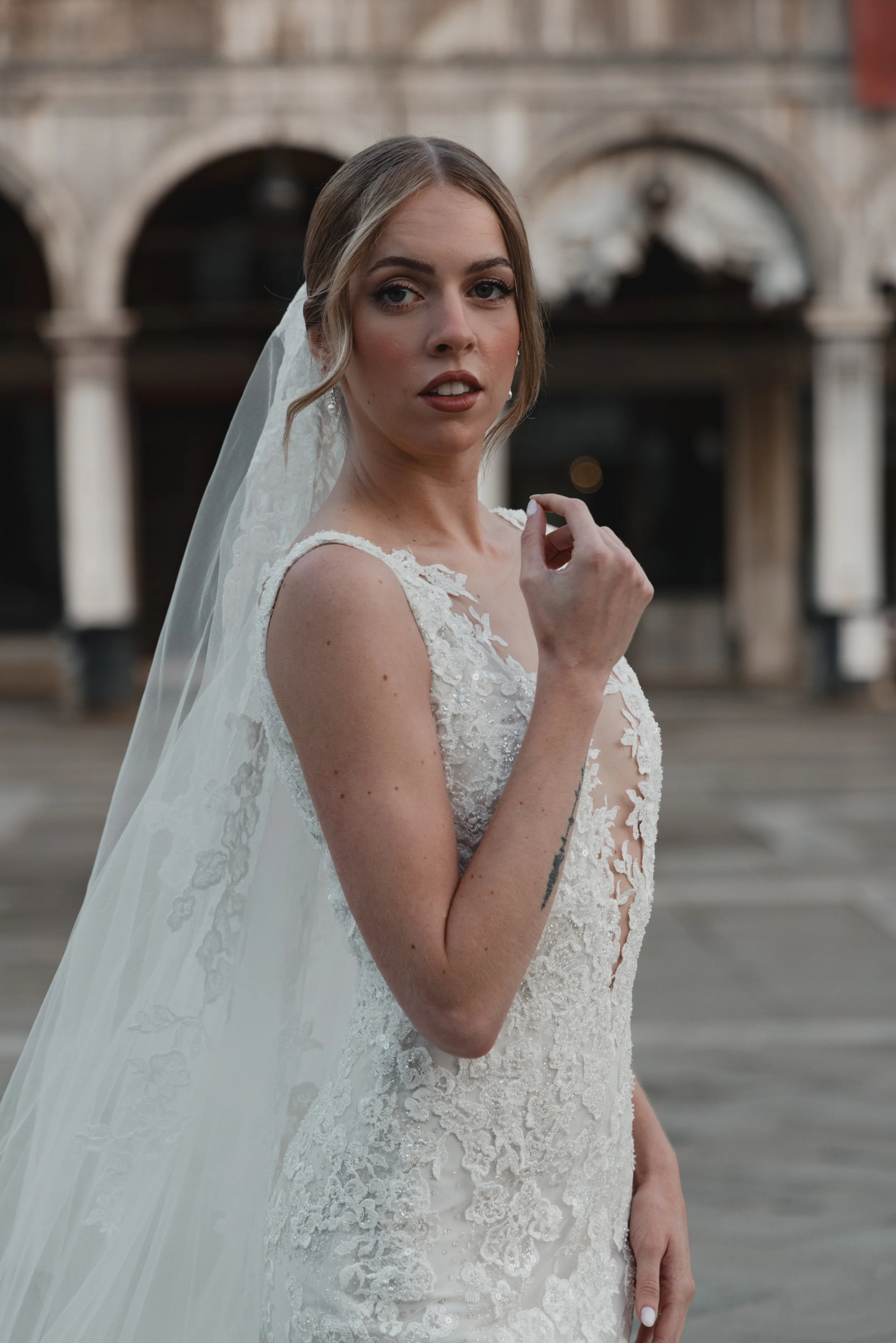 A woman wearing a wedding dress with lace details and a veil, standing outdoors with an architectural background featuring arches and columns.