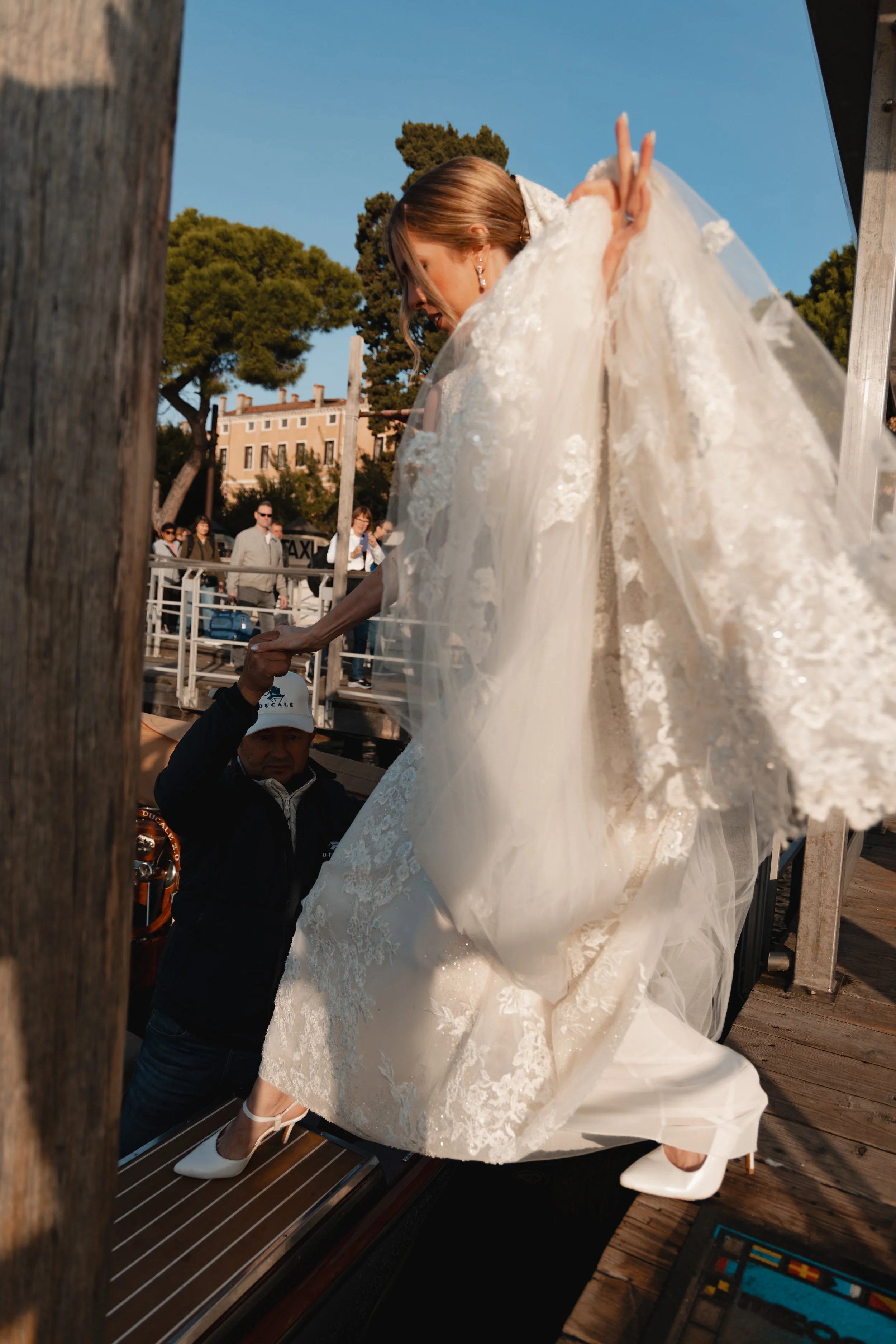A bride in a white lace wedding dress and white heels is stepping onto a boat, assisted by a man, on a wooden dock. The scene is outdoors during the daytime with a blue sky and green trees in the background, and other people are visible in the backgr
