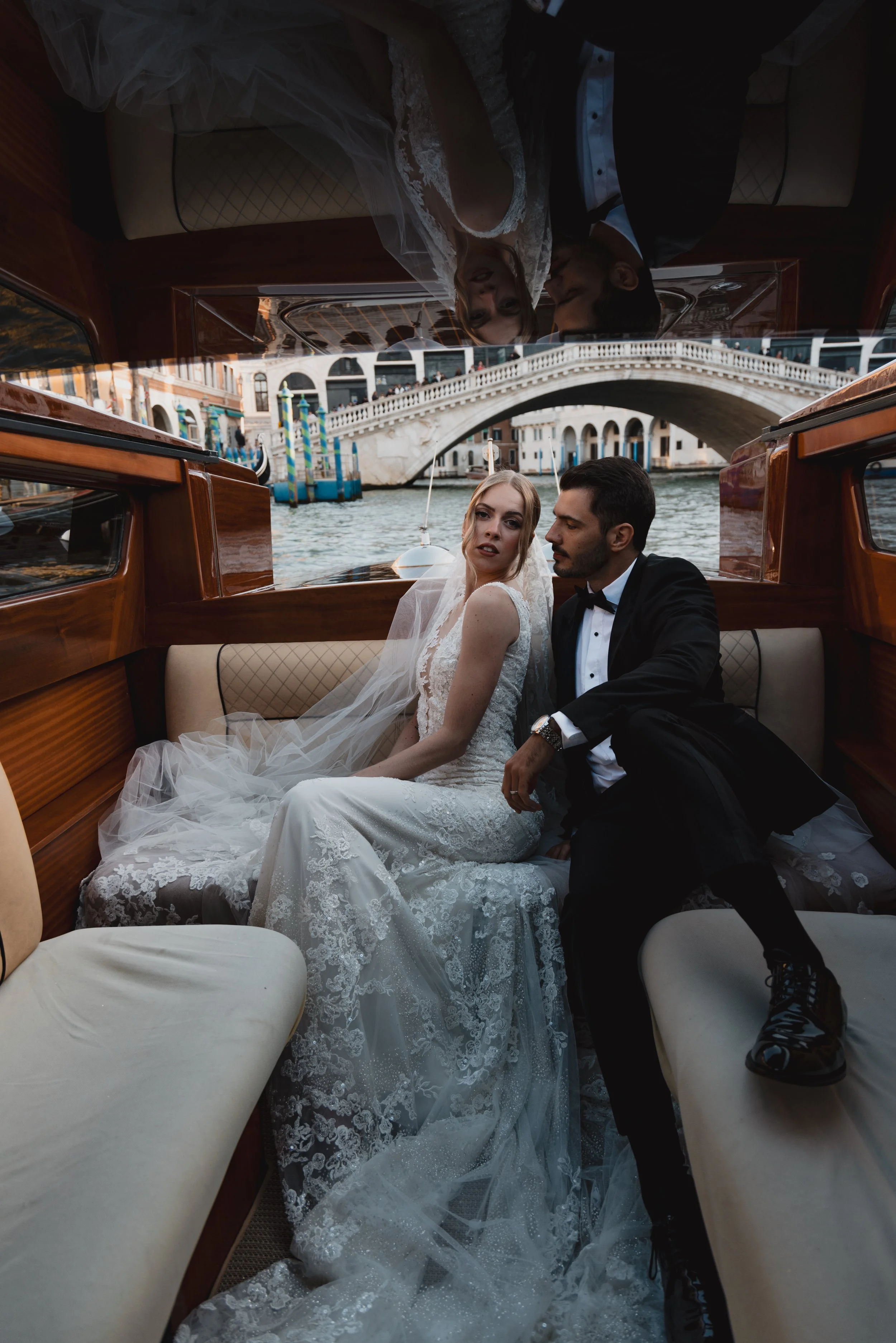 A bride and groom sitting inside a boat on a canal in Venice, Italy. The bride is wearing a white wedding dress with lace details, and the groom is dressed in a black tuxedo. The background shows the iconic Rialto Bridge and historic buildings along 