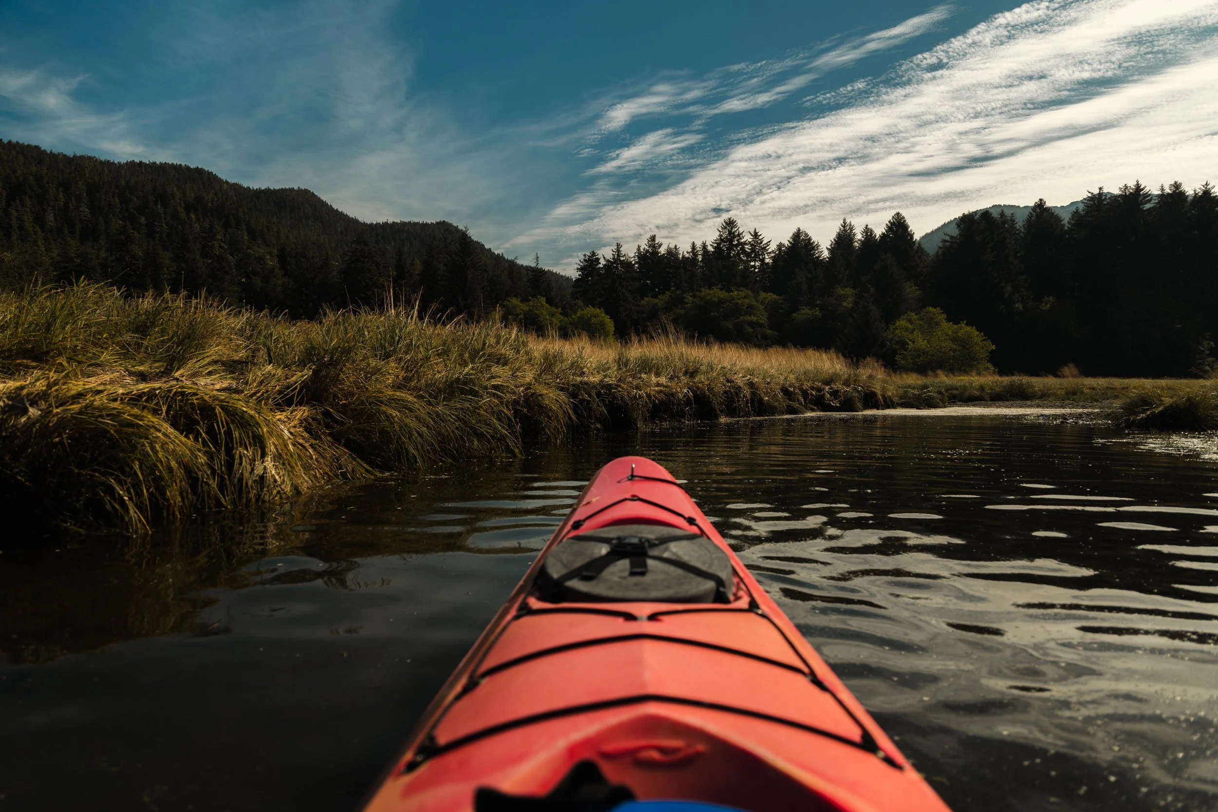 View from a kayak on a river surrounded by trees and mountains with a partly cloudy sky.