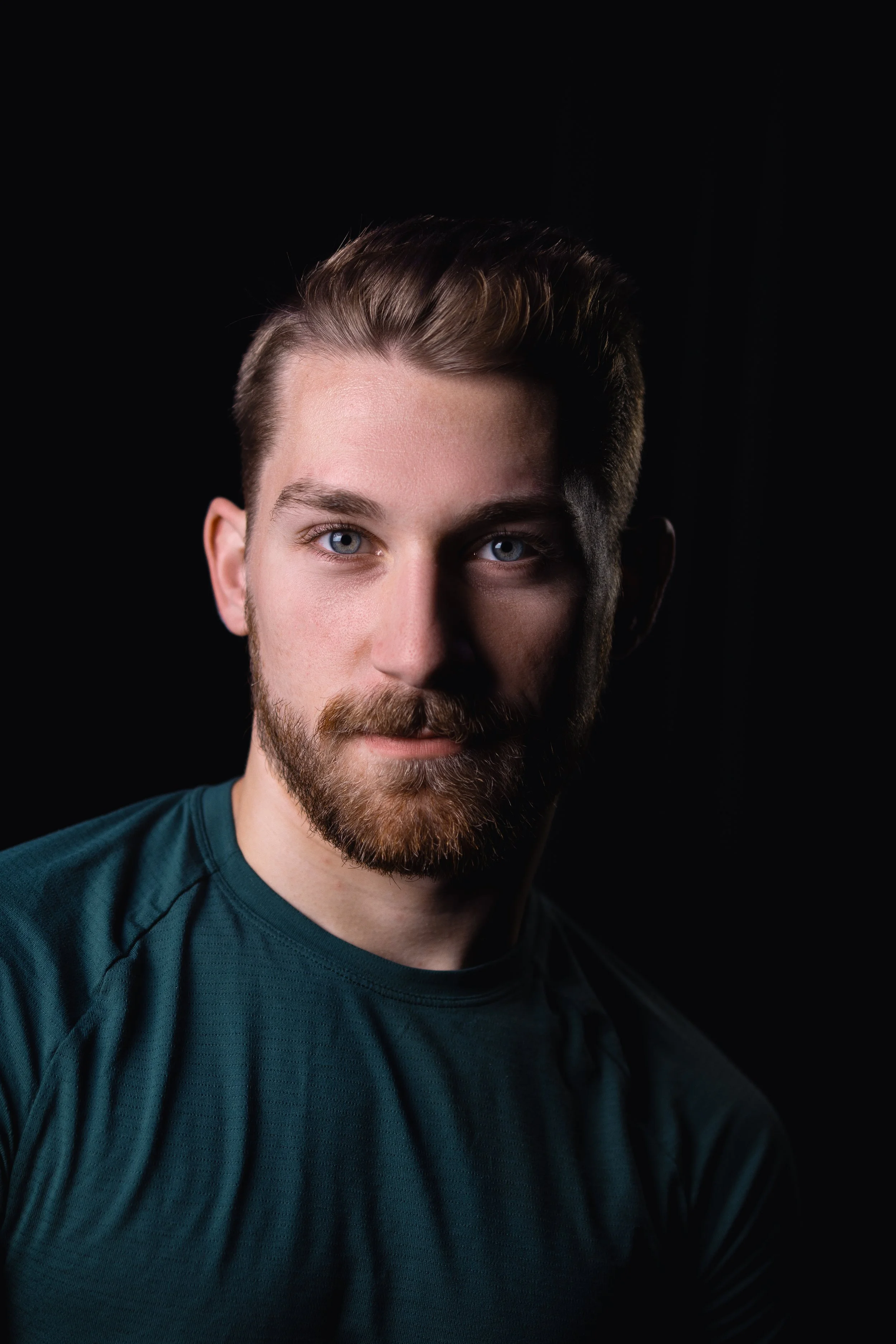Close-up of a young man with brown hair, blue eyes, and a beard, wearing a dark teal athletic shirt, against a black background.