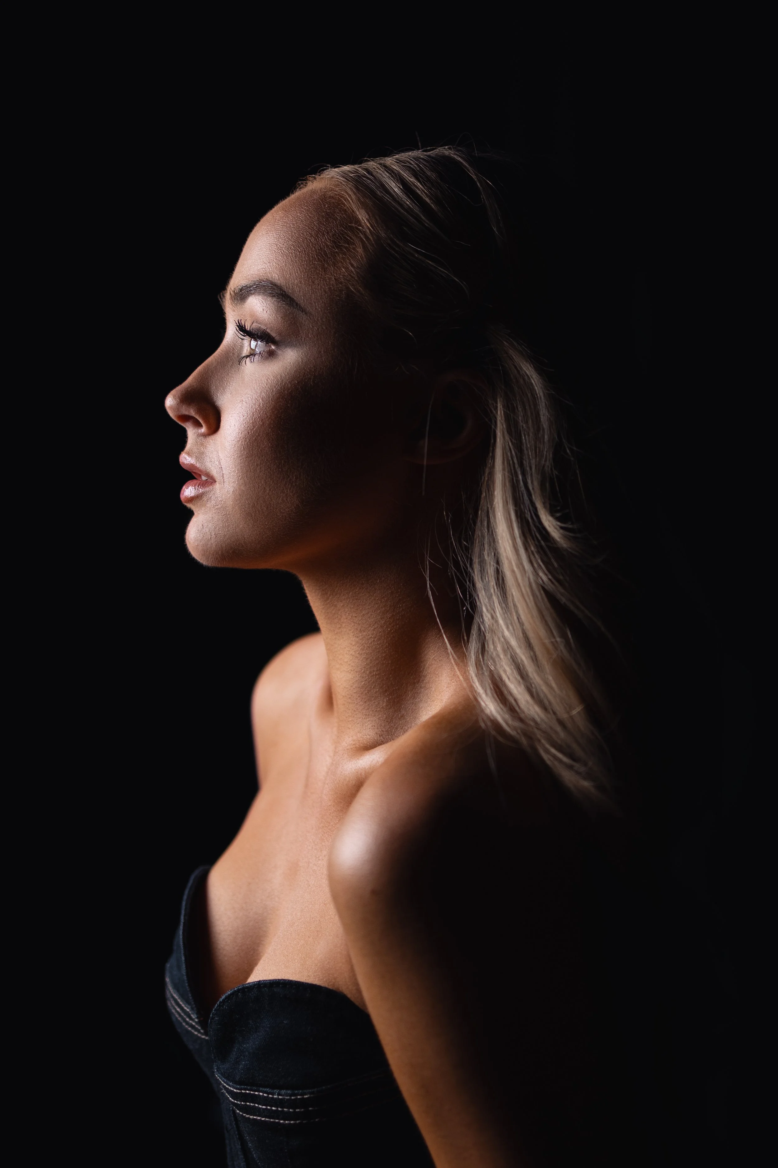 A side profile portrait of a woman with light skin and long blonde hair, looking upward, against a black background.