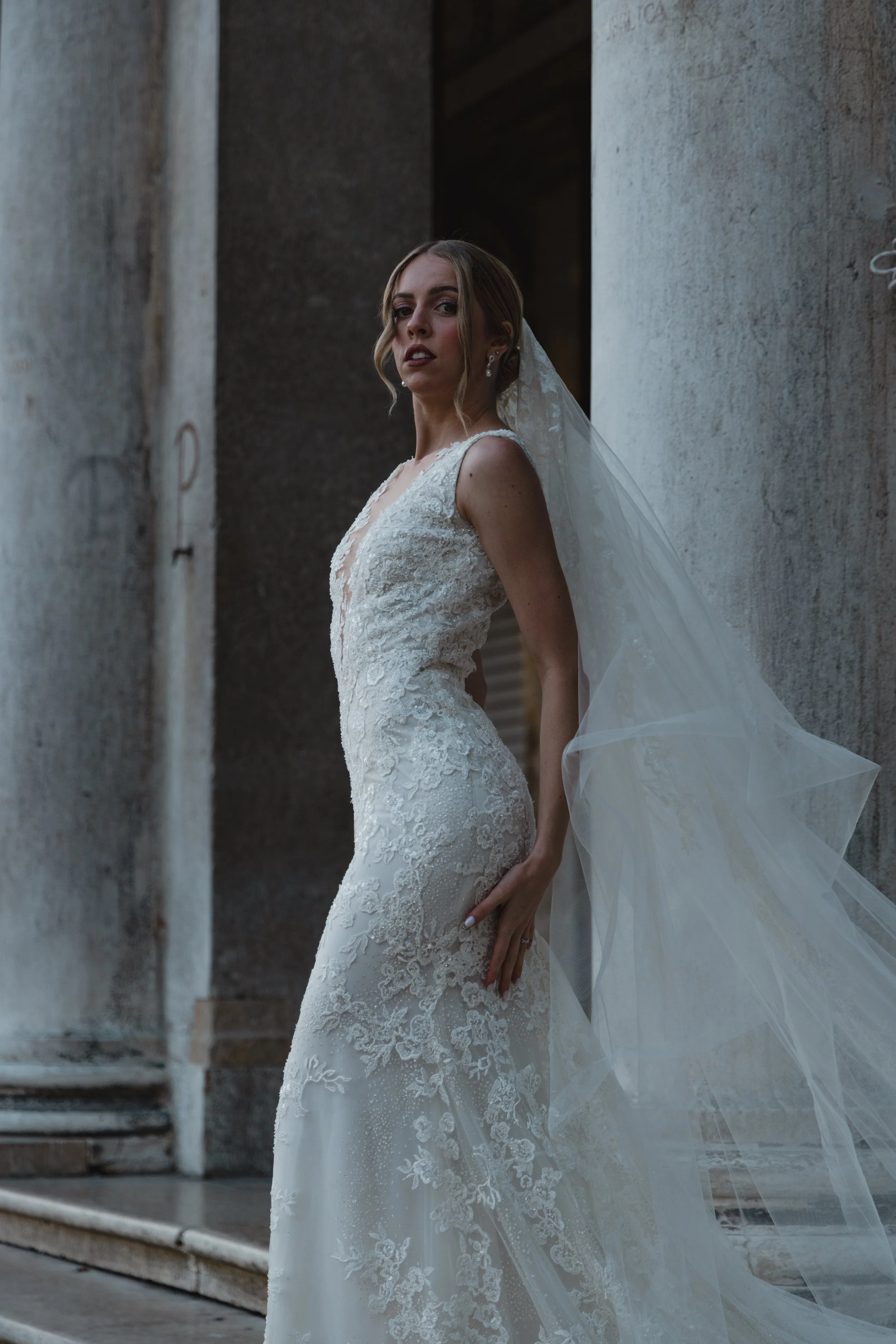 A bride in a white lace wedding gown standing outside between two concrete columns.