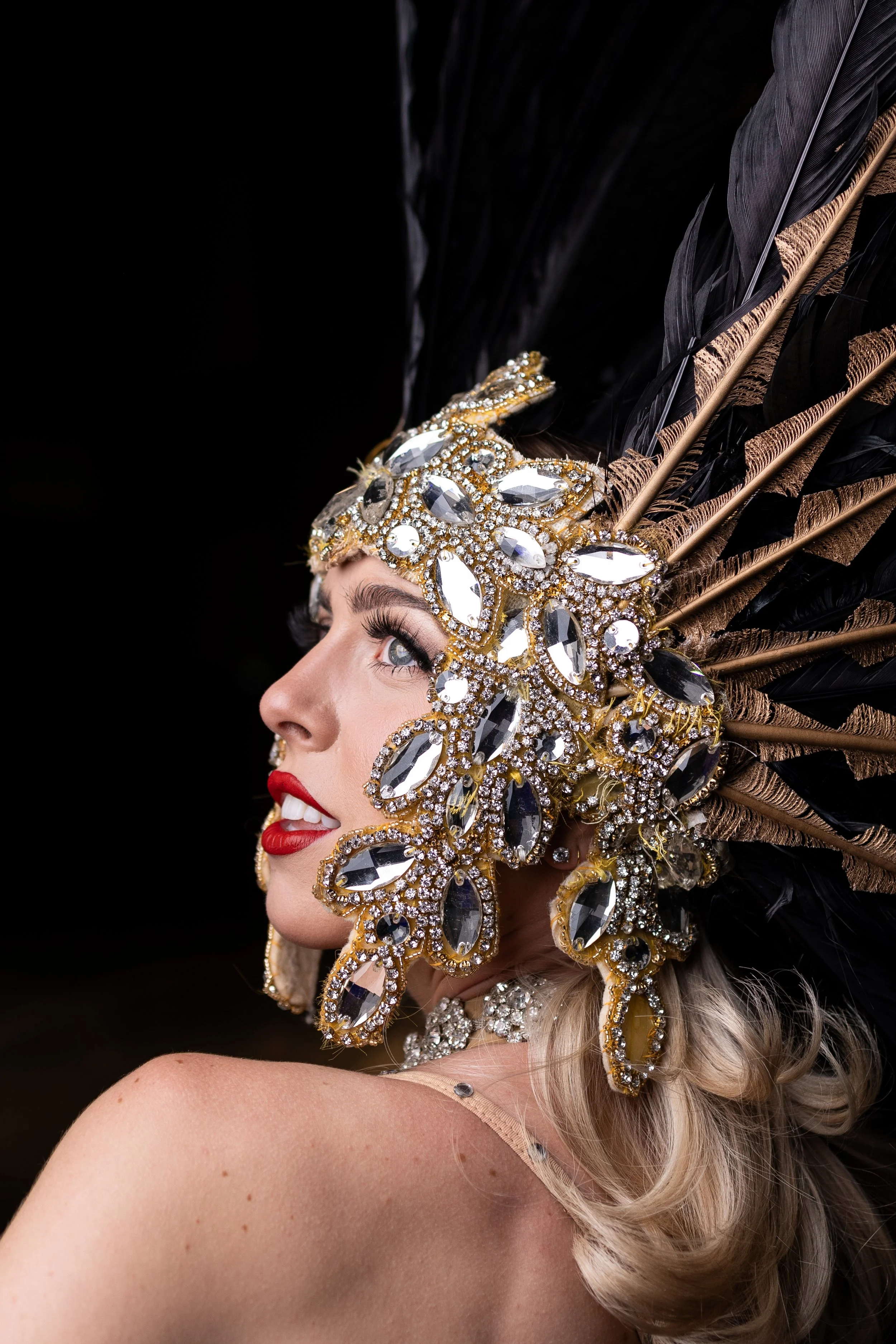 Close-up of a woman wearing a large, elaborate headdress decorated with black feathers, gold accents, and large crystal-like gemstones. She has bright red lipstick, long eyelashes, and blonde hair styled in loose waves.