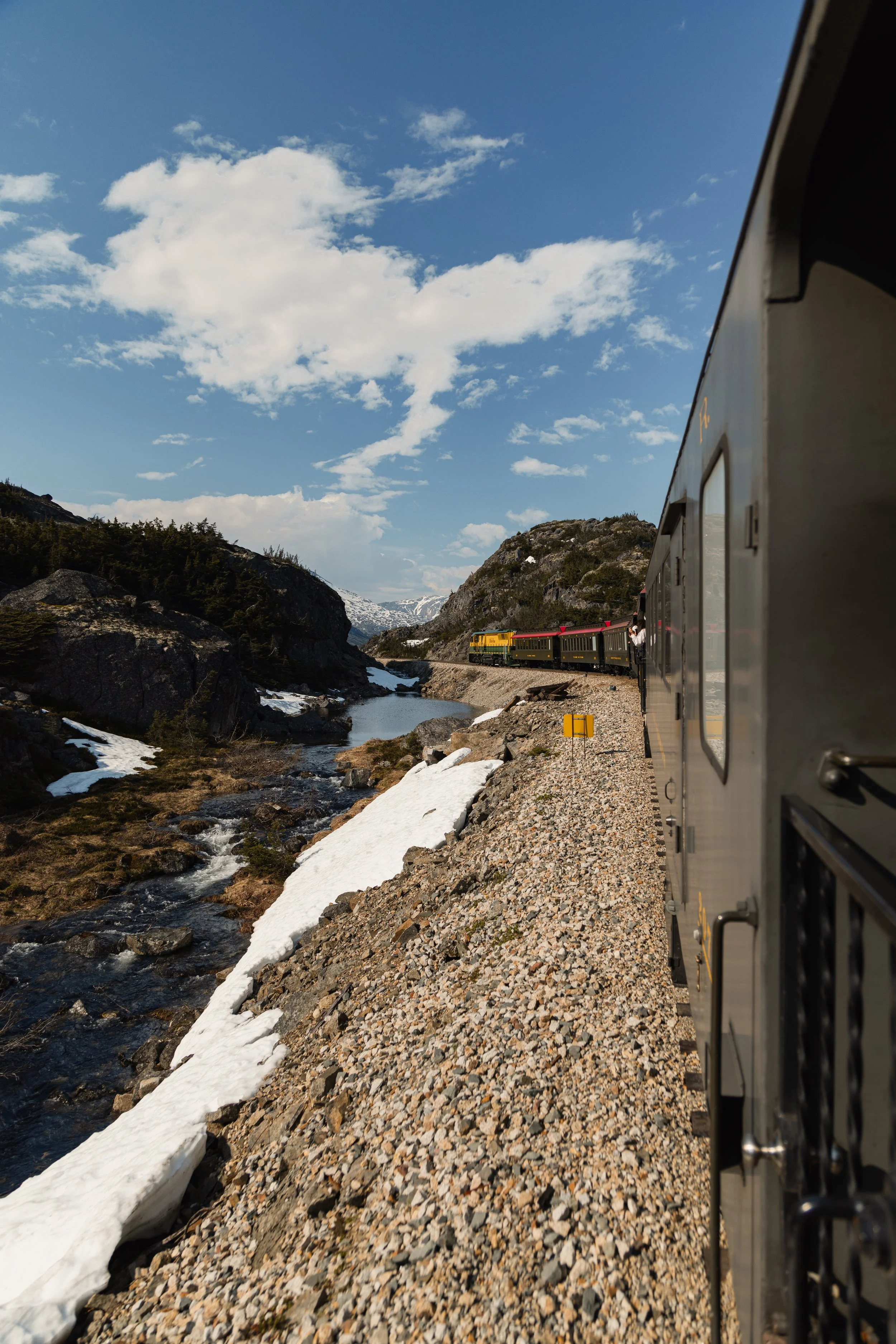 View from a train window showing a rocky landscape with a small stream, patches of snow, green trees on hills, a mountainous backdrop, blue sky with scattered clouds, and the side of a train car in the foreground.