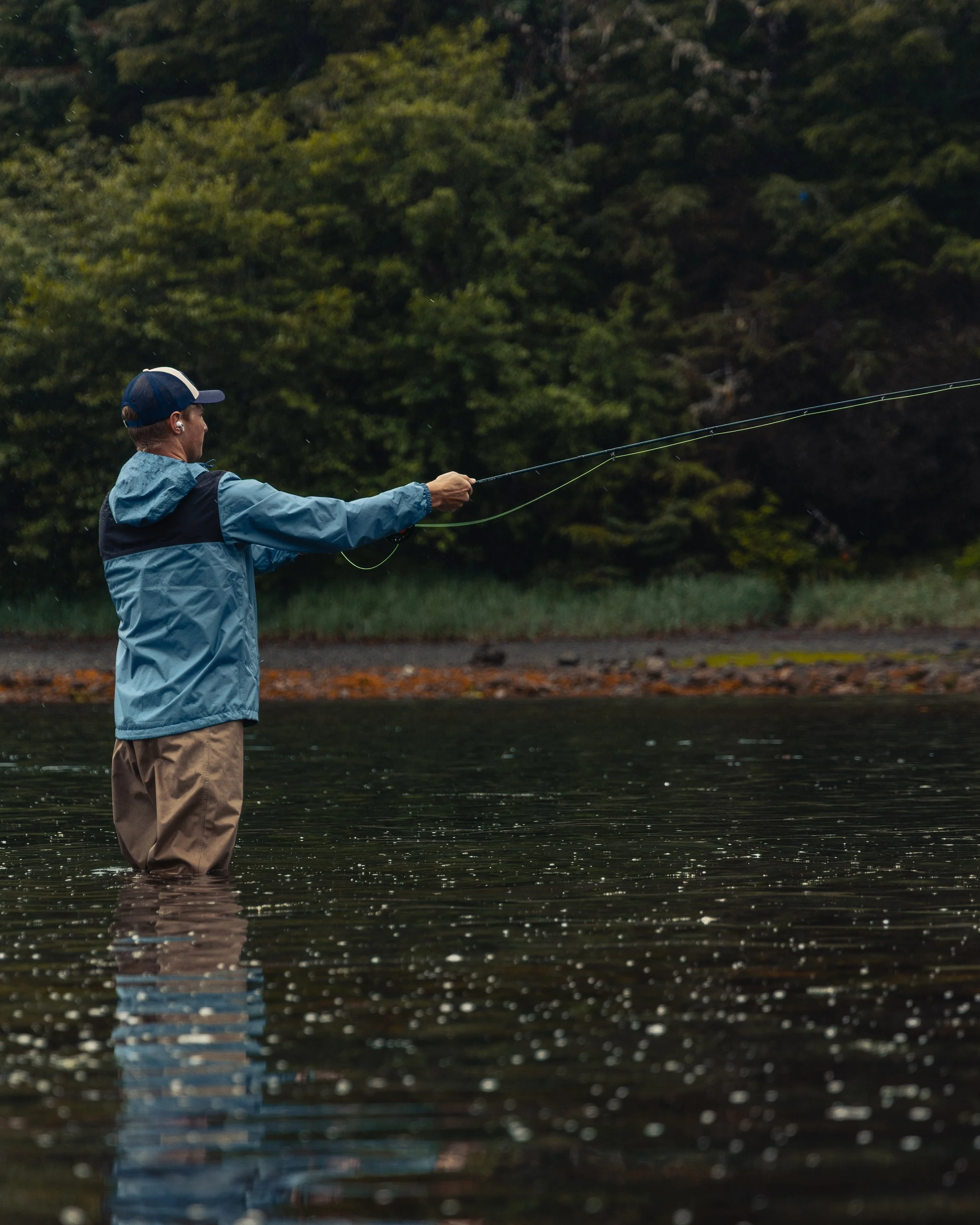 A person standing in a river, fly fishing, wearing a blue jacket, khaki pants, and a blue cap, with a dense forest in the background.