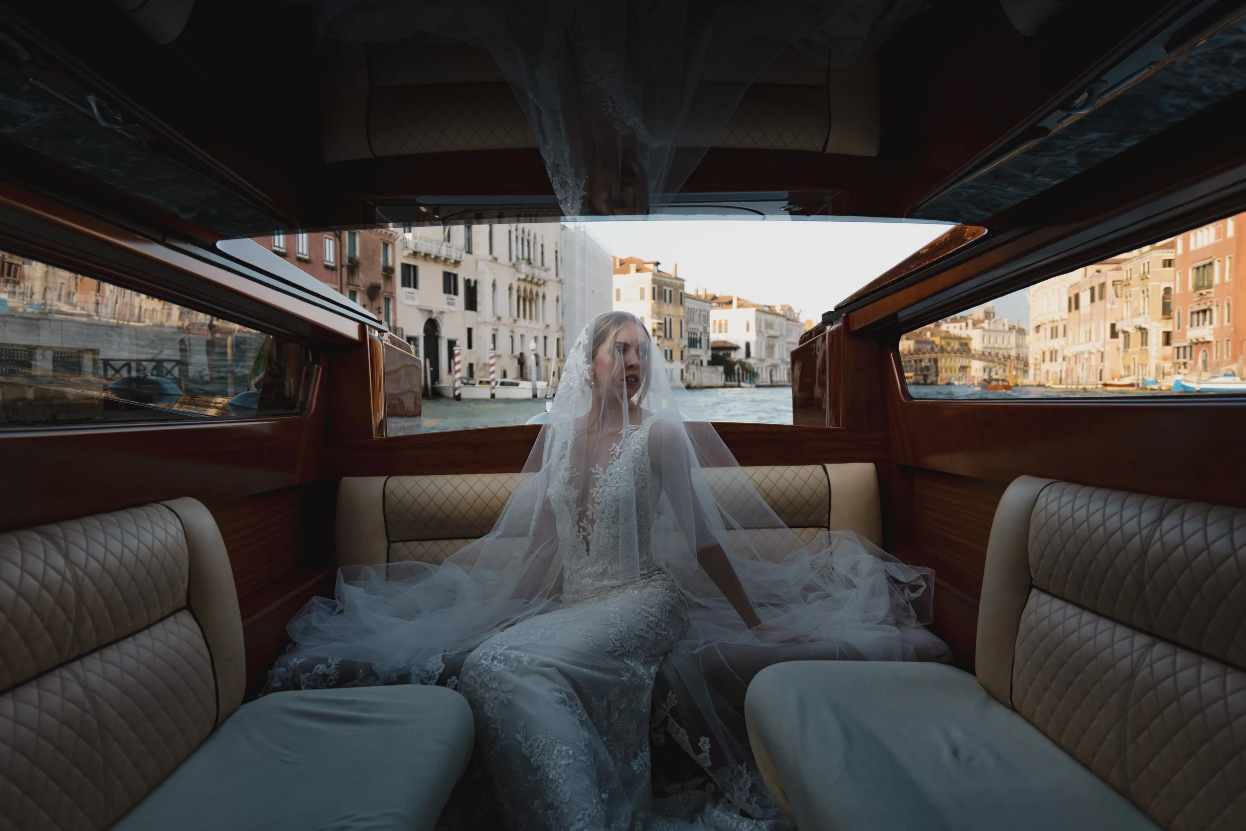 A woman in a wedding dress sitting inside a boat with large windows, overlooking colorful buildings along a canal in Venice, Italy.
