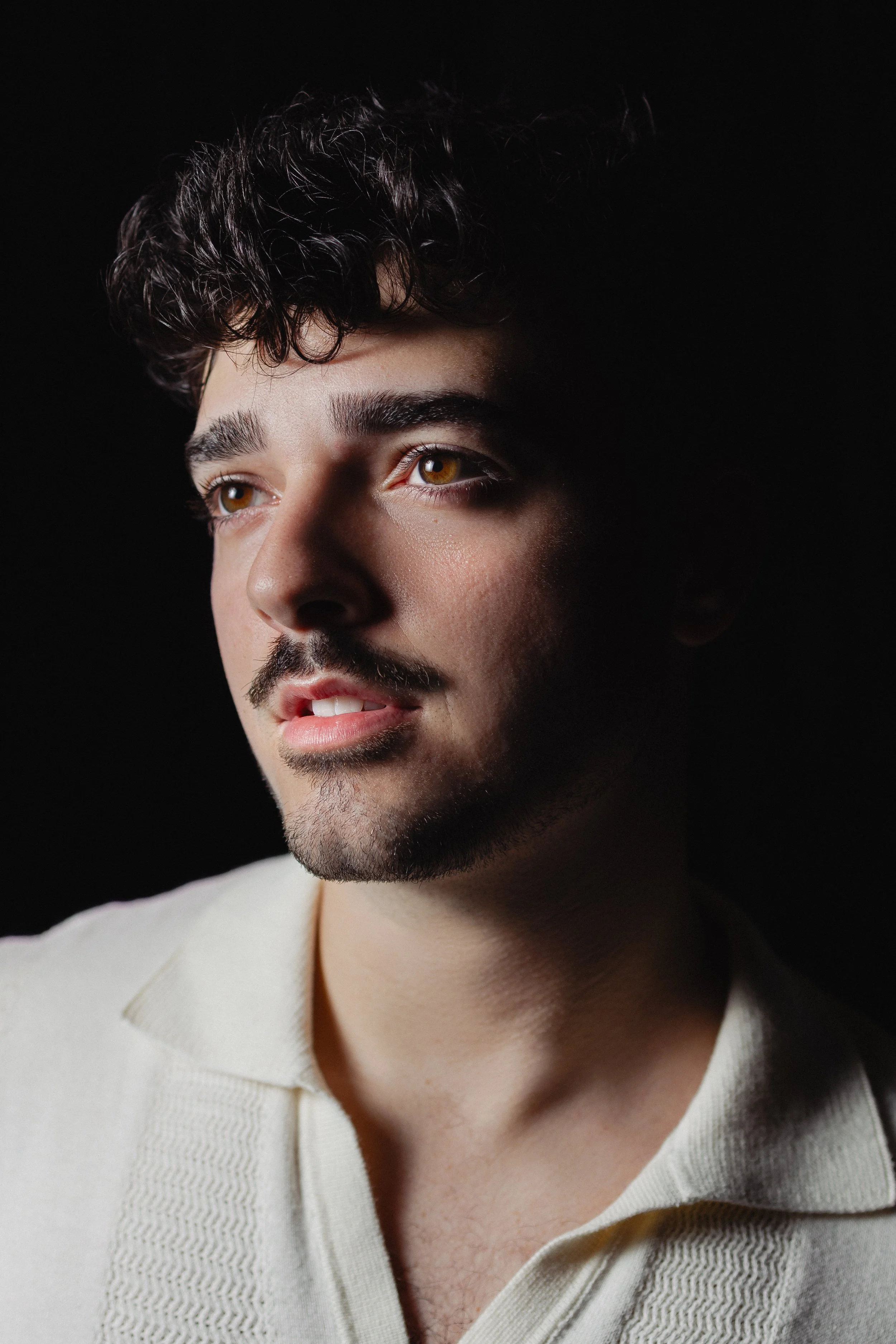 Close-up portrait of a young man with curly dark hair, hazel eyes, and a light beard, wearing a cream-colored sweater against a black background.