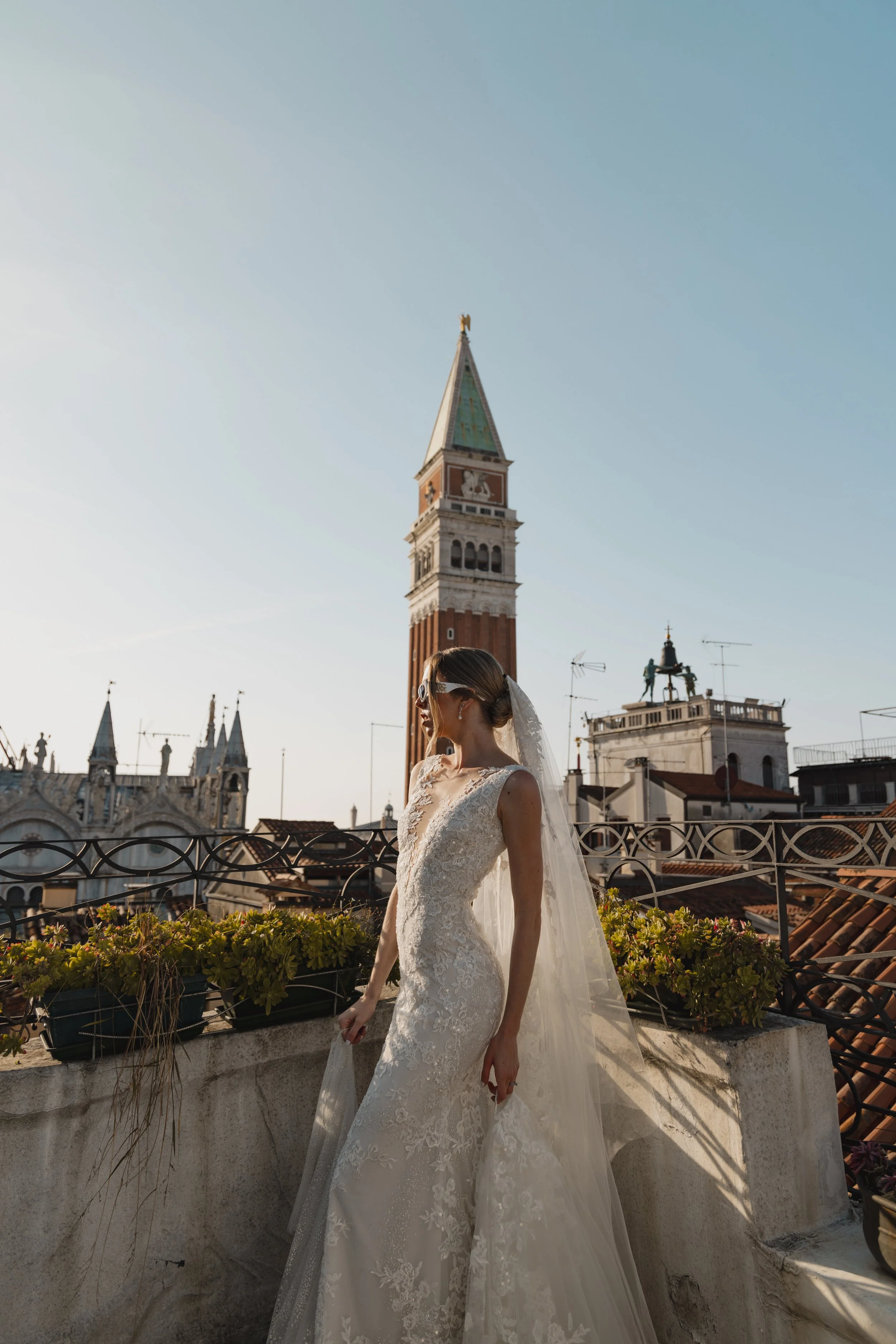 A bride in a lace wedding dress and veil standing on a rooftop with historic Venetian architecture and the clock tower in Venice, Italy, in the background.