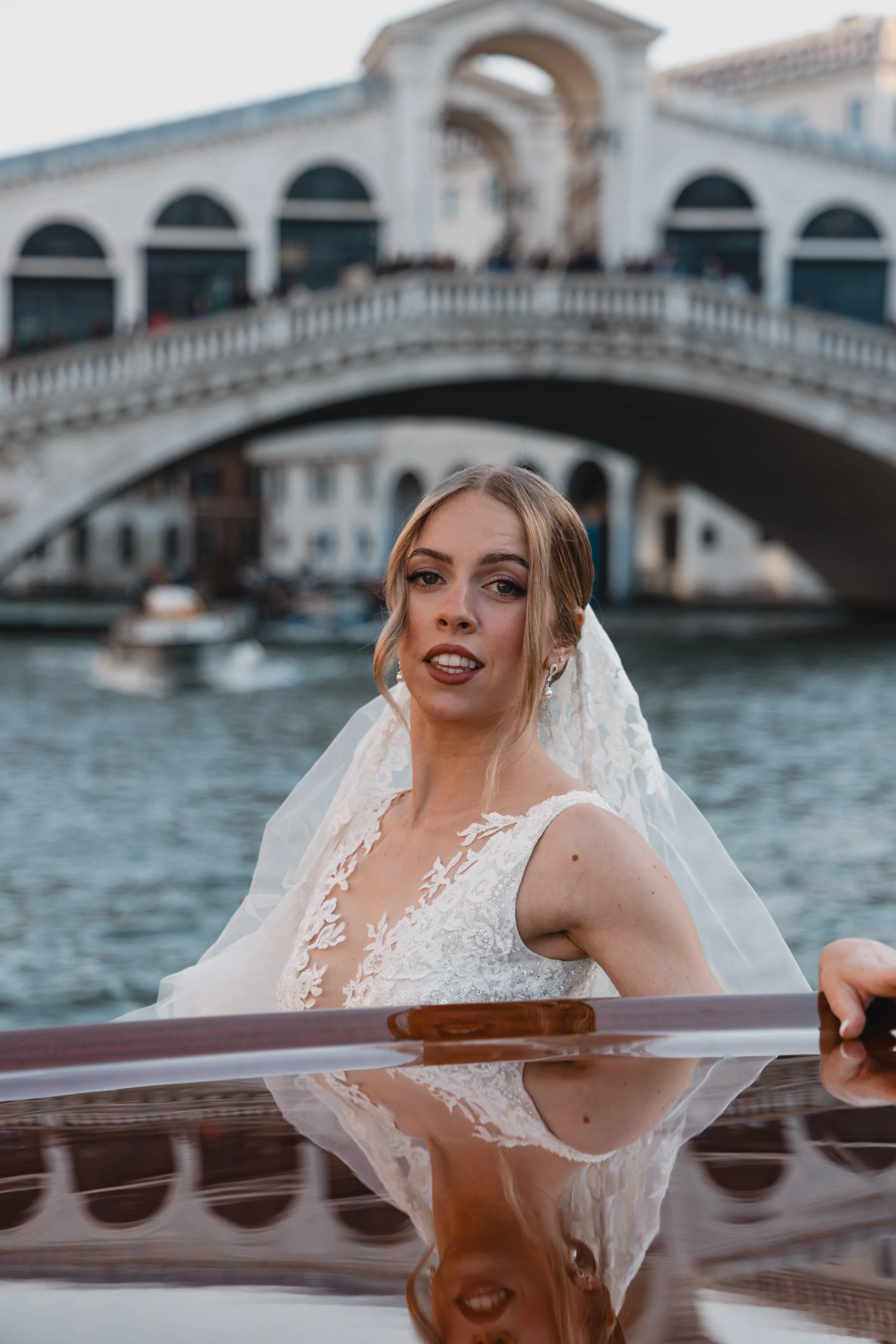A woman in a wedding dress on a boat with the city of Venice and a bridge in the background.