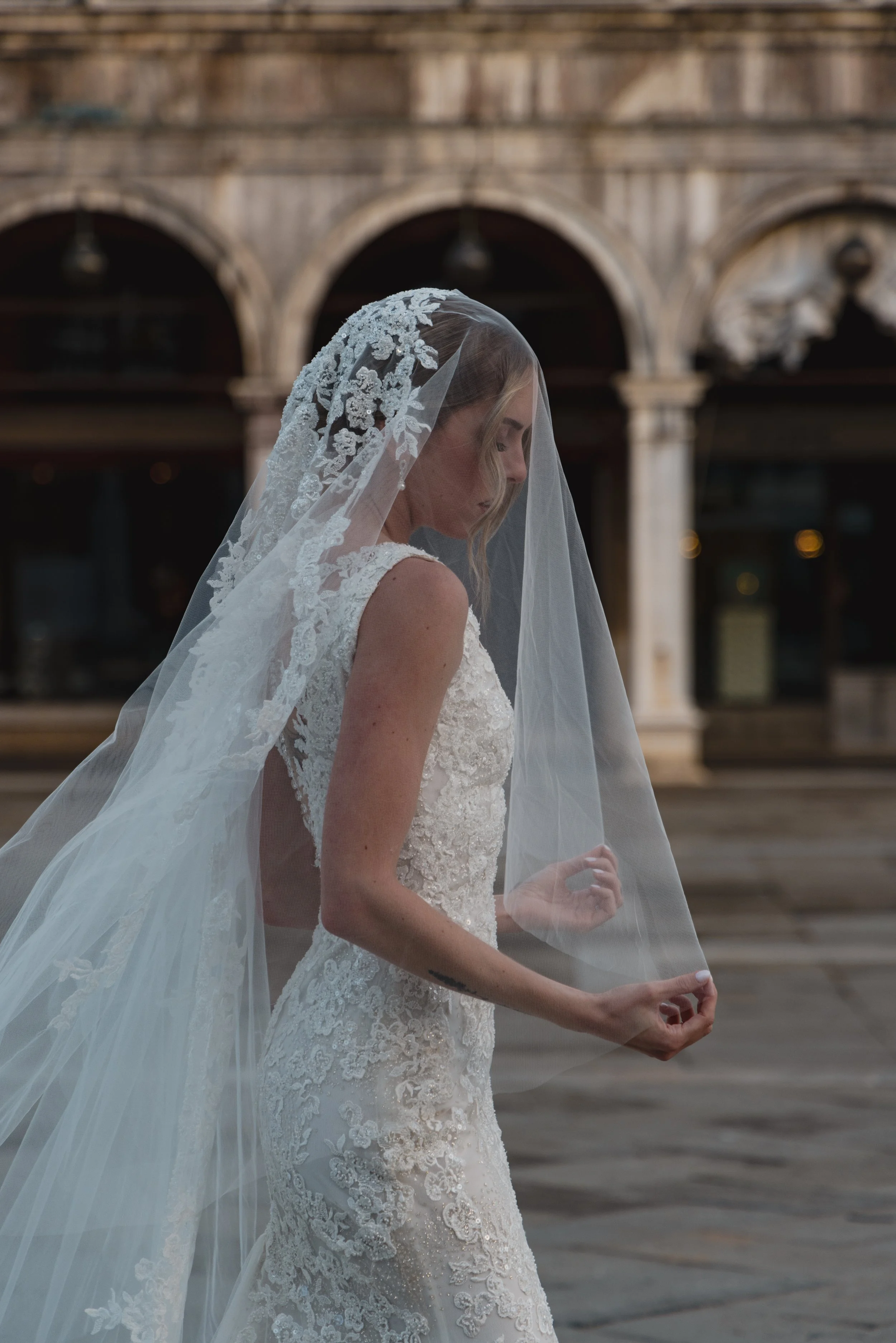 A woman in a white bridal gown and lace veil stands outdoors near an arched stone building, looking down with her eyes closed.