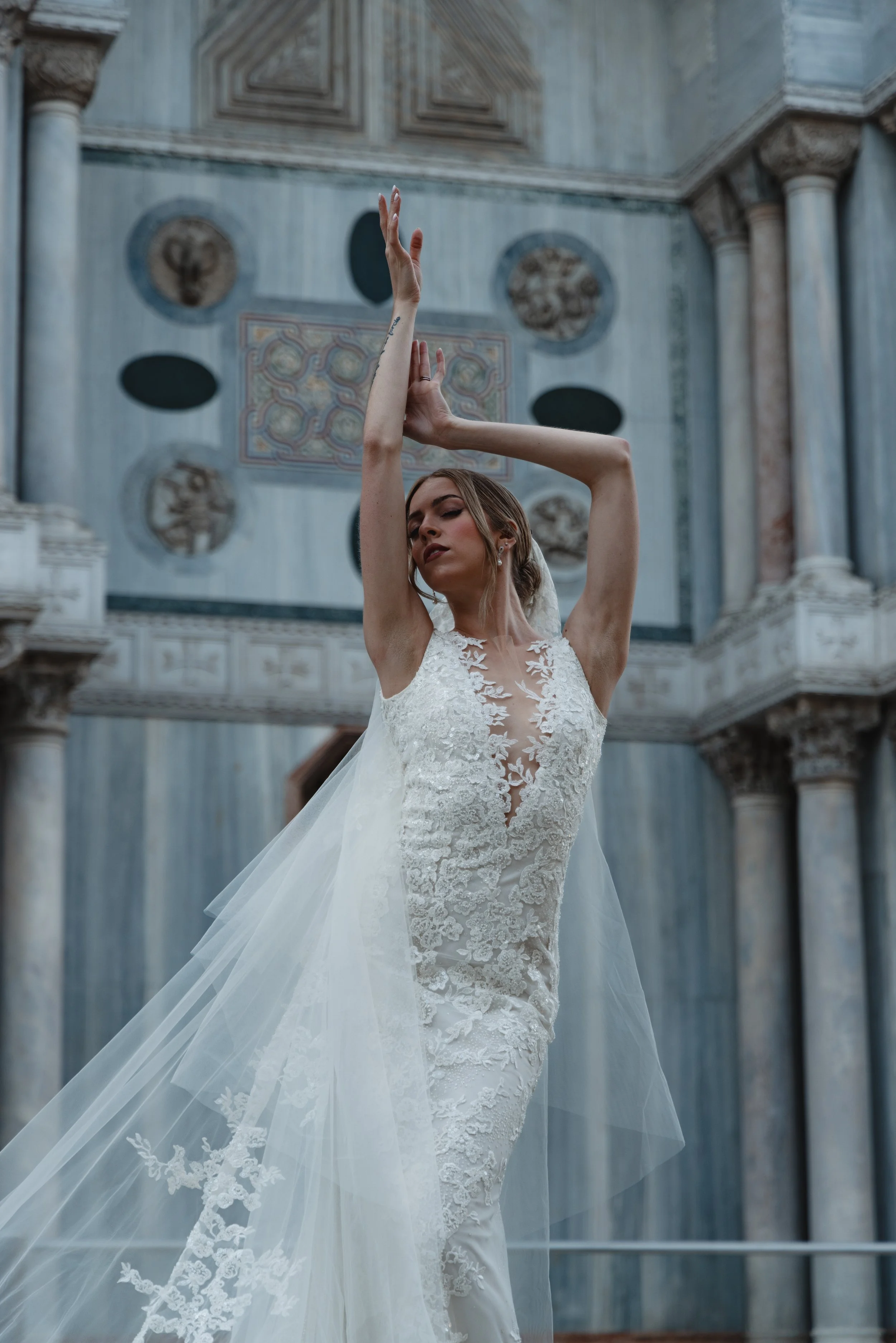 A woman in a wedding dress dancing or posing inside a grand, historic building with marble columns and decorative ceiling details.