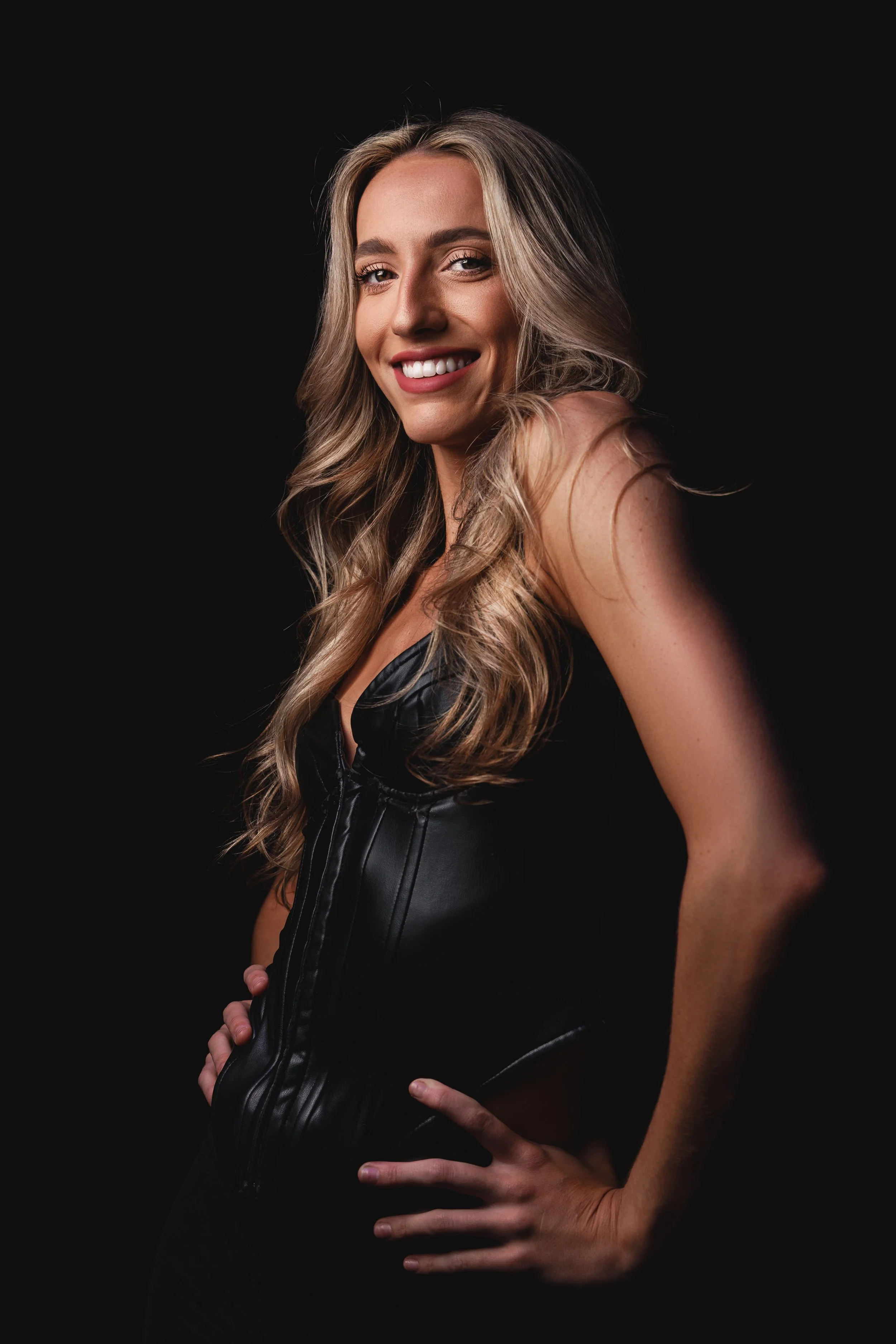 A woman with long blonde hair, smiling, wearing a black leather outfit, against a black background.