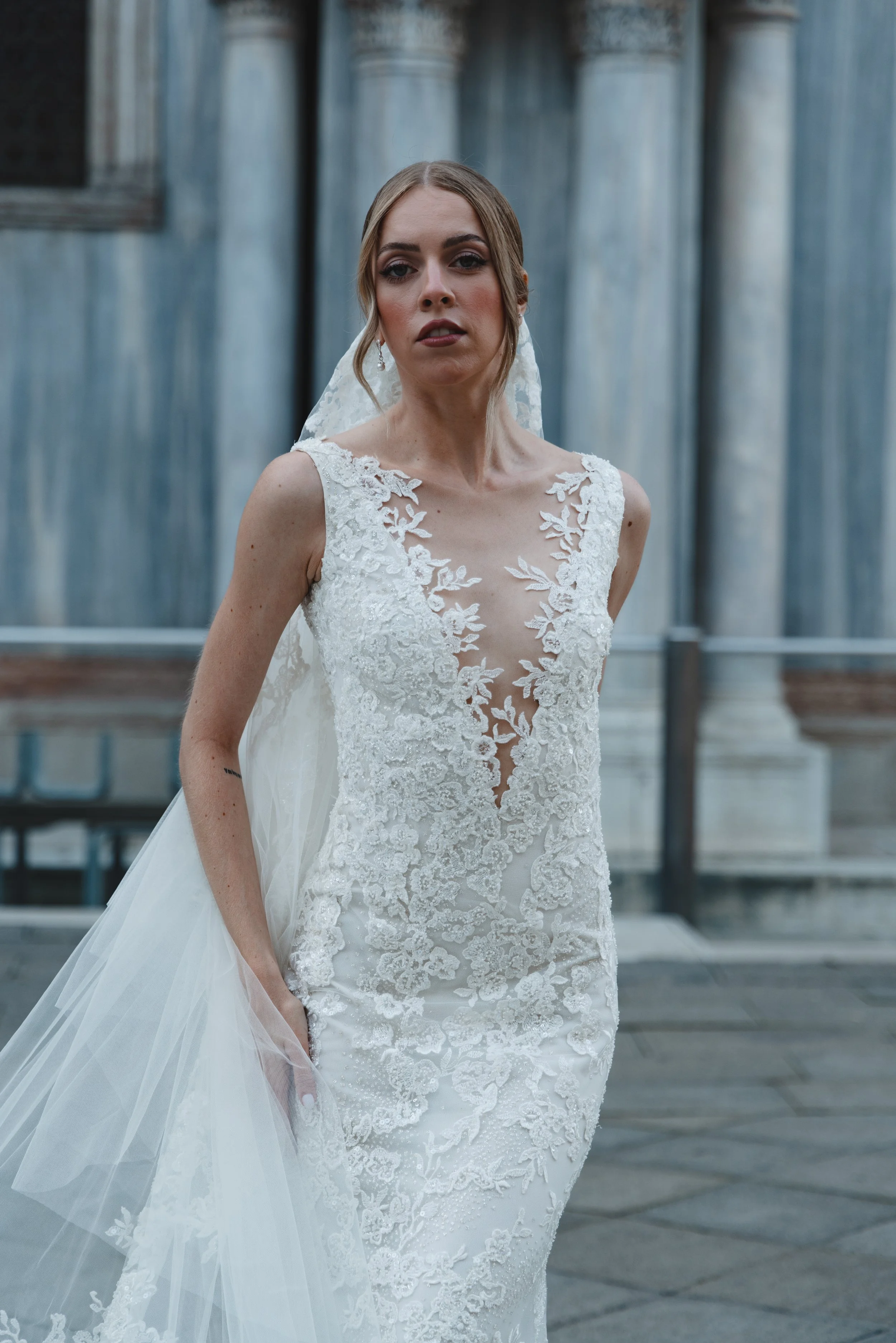 A woman in an elegant white wedding dress with lace detailing, standing outdoors in front of large metallic columns.
