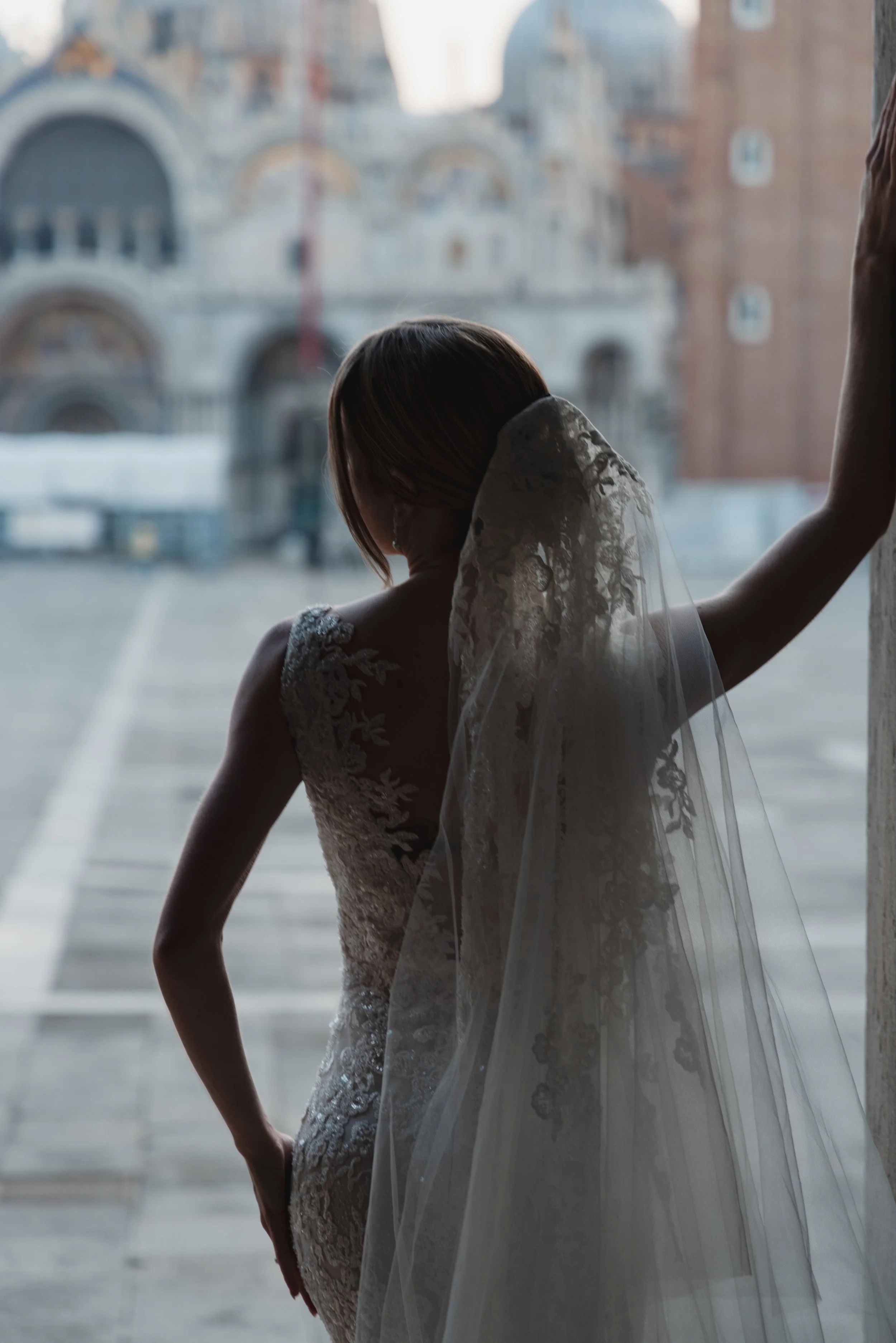 Bride standing by a window in a wedding dress with lace details, looking down, with a building visible outside the window.