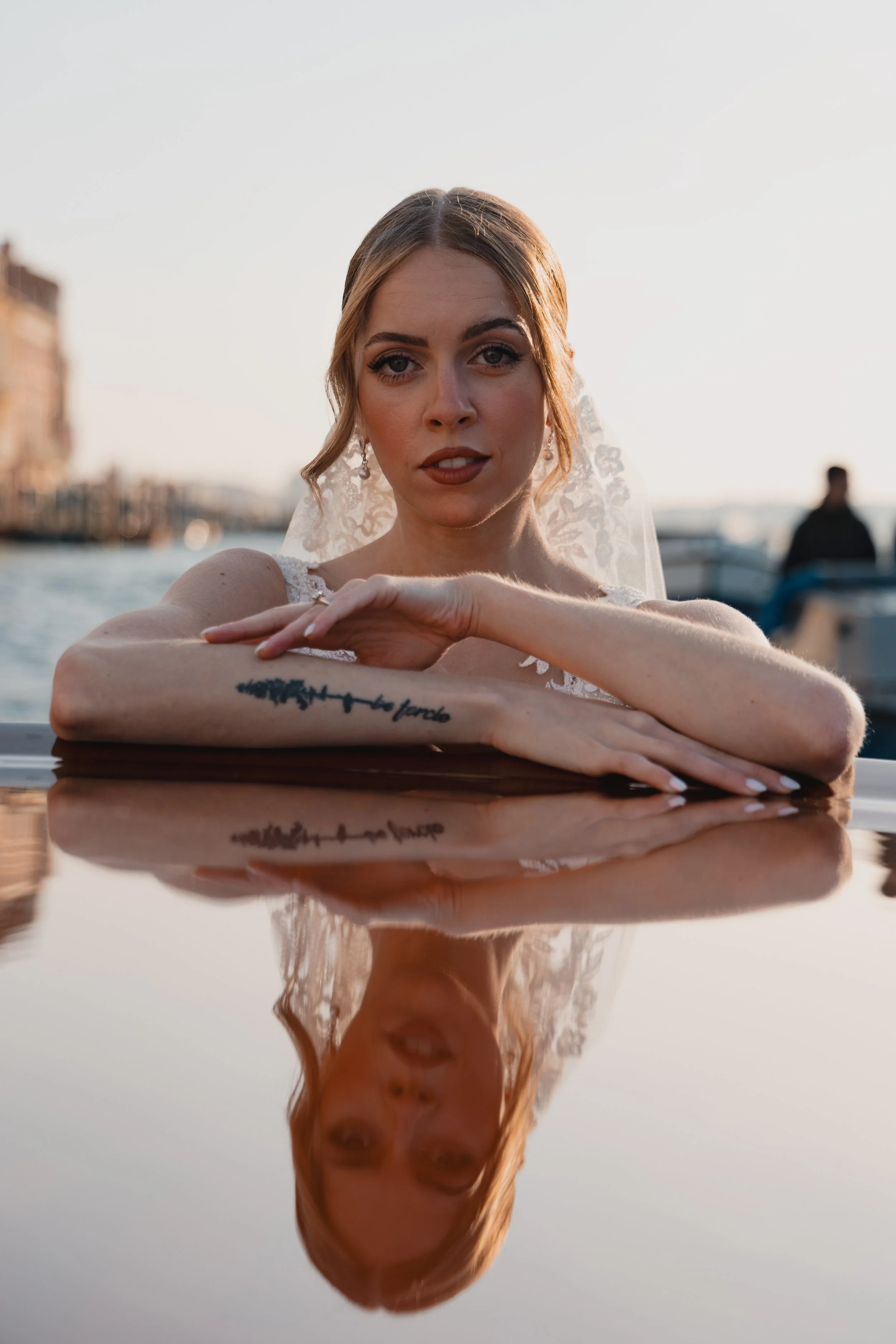 A woman wearing a wedding dress and veil is leaning on a reflective water surface, with her reflection visible. She has light brown hair, blue eyes, and a tattoo on her forearm that reads 'la forza'. The background features a blurry cityscape with bu
