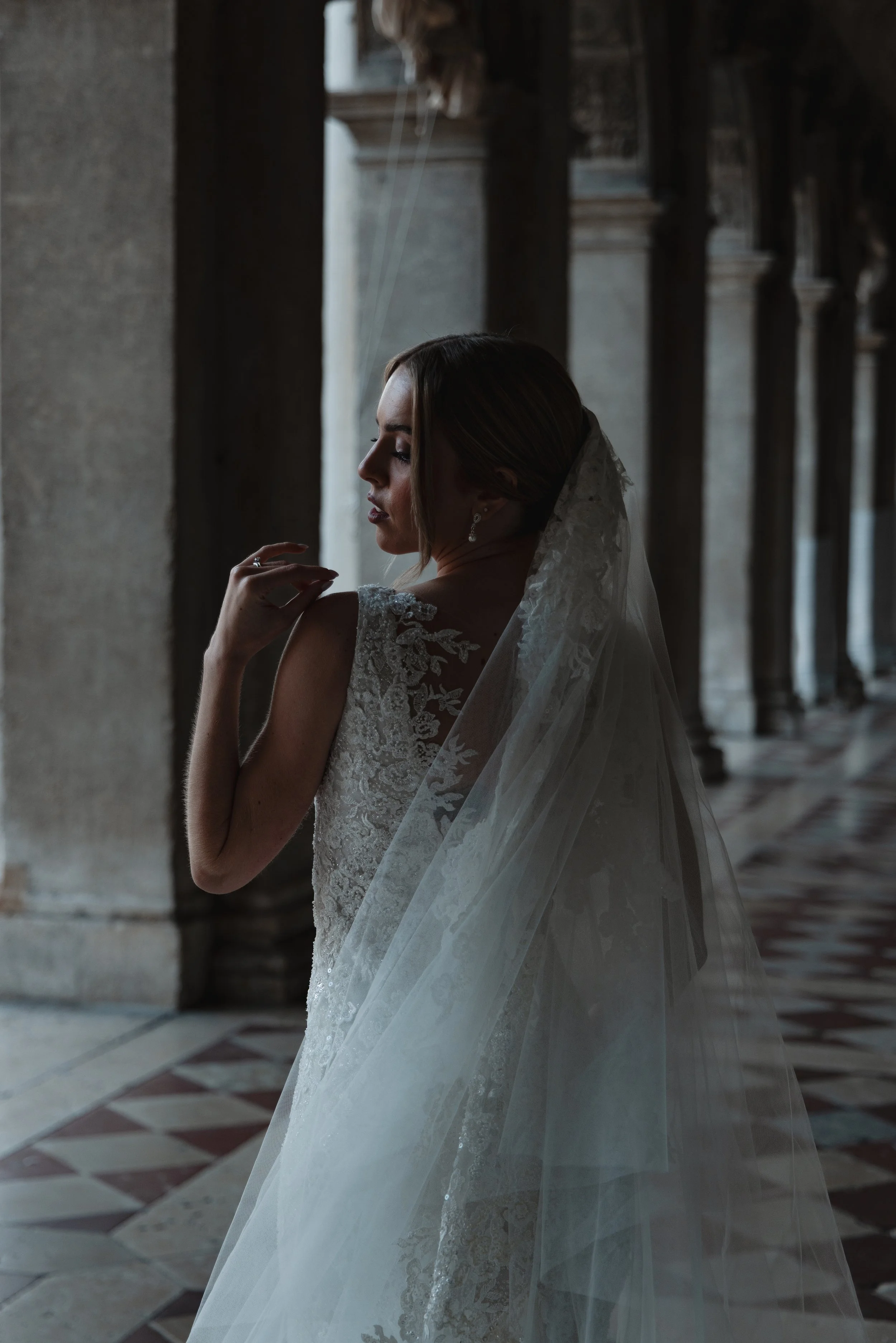 A bride in a lace wedding gown with a veil, standing in a historic stone corridor with arches and patterned tile flooring.