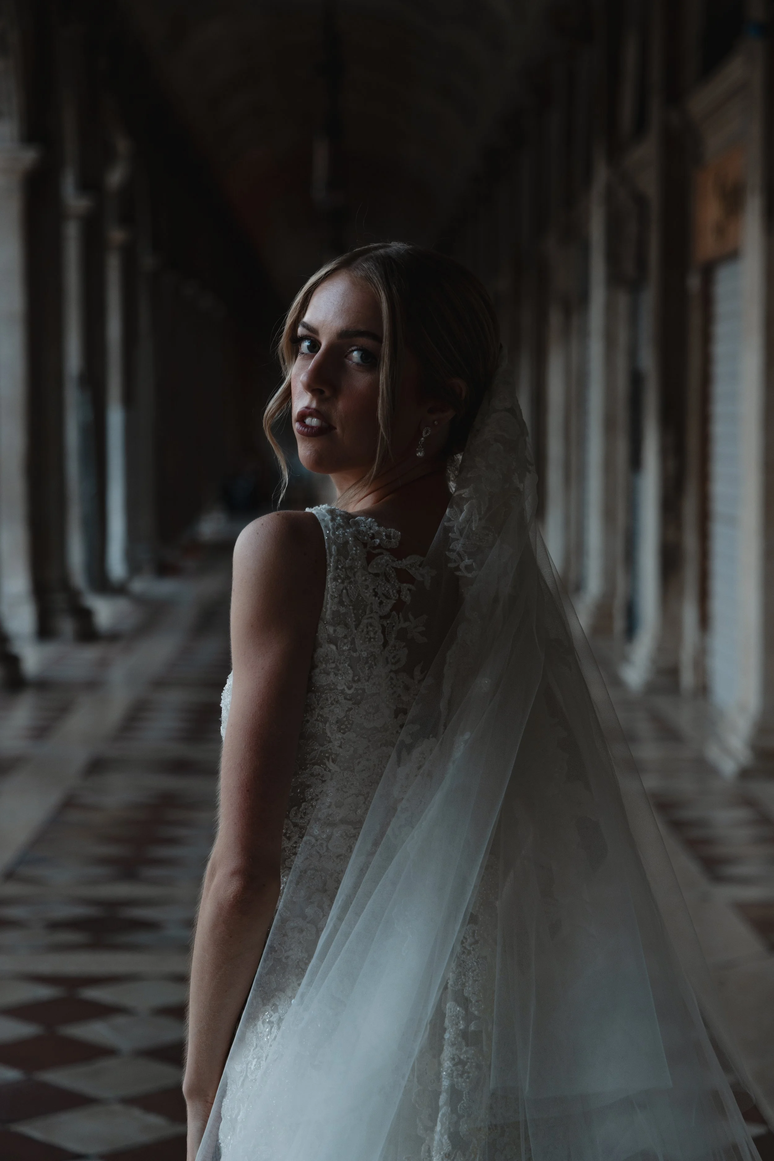 A bride in a lace wedding dress with a veil standing in a dimly lit corridor with patterned flooring and ornate walls.