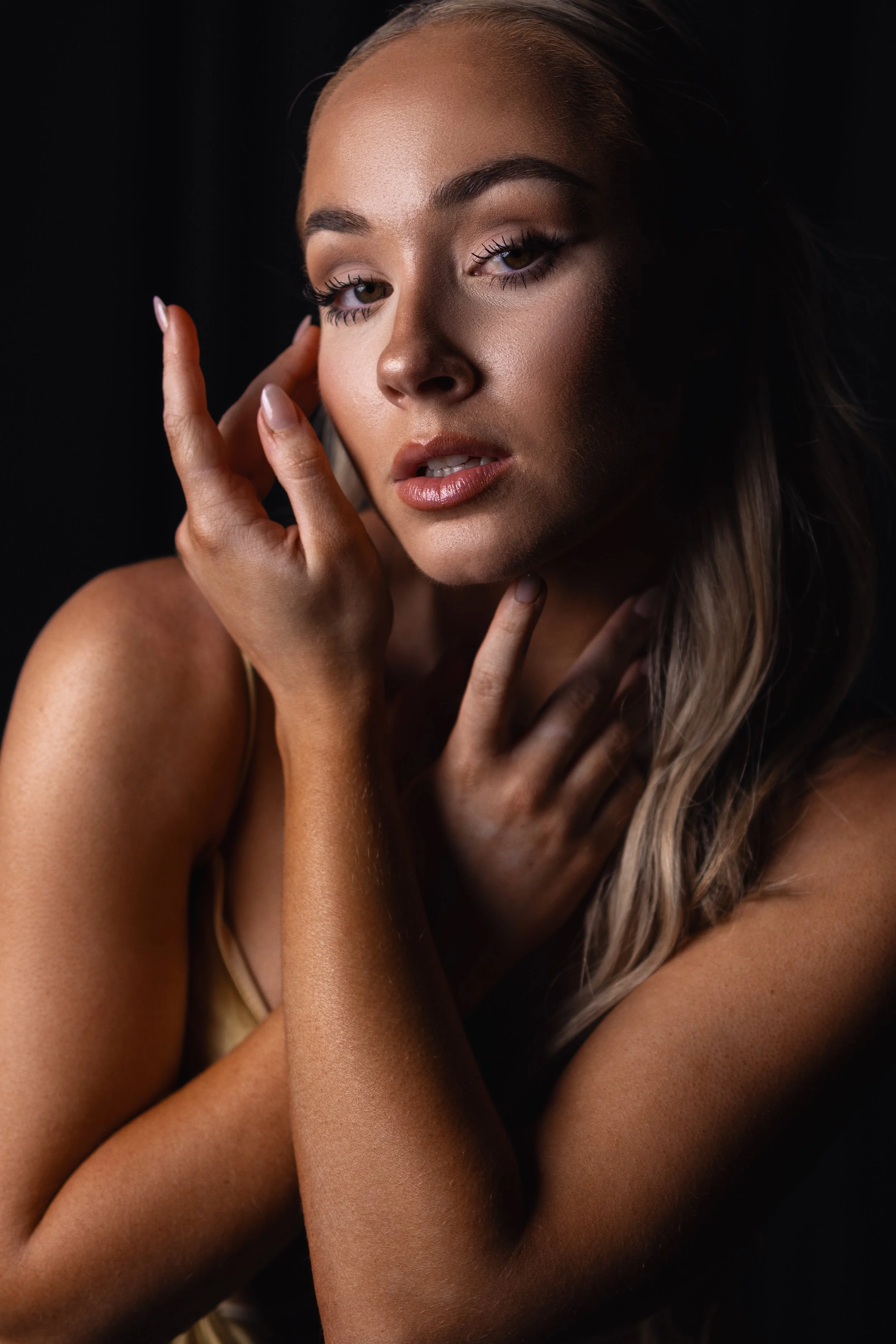 Close-up of a woman with light skin, brown hair, and brown eyes, touching her face gently with her fingers against a dark background.