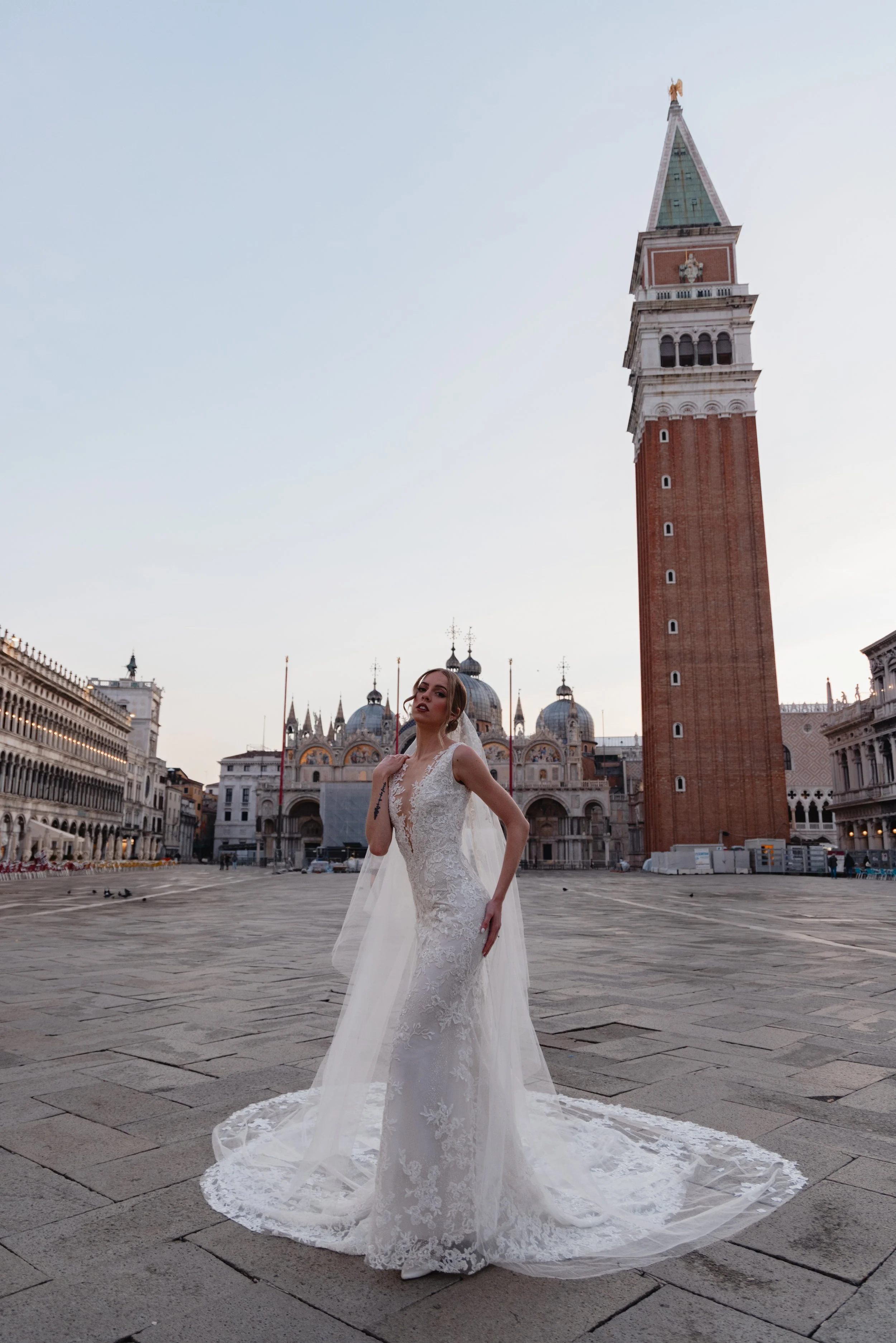 A woman in a white lace wedding dress standing in St. Mark's Square in Venice, Italy, with the St. Mark's Campanile tower and domed basilica visible in the background, during dusk.
