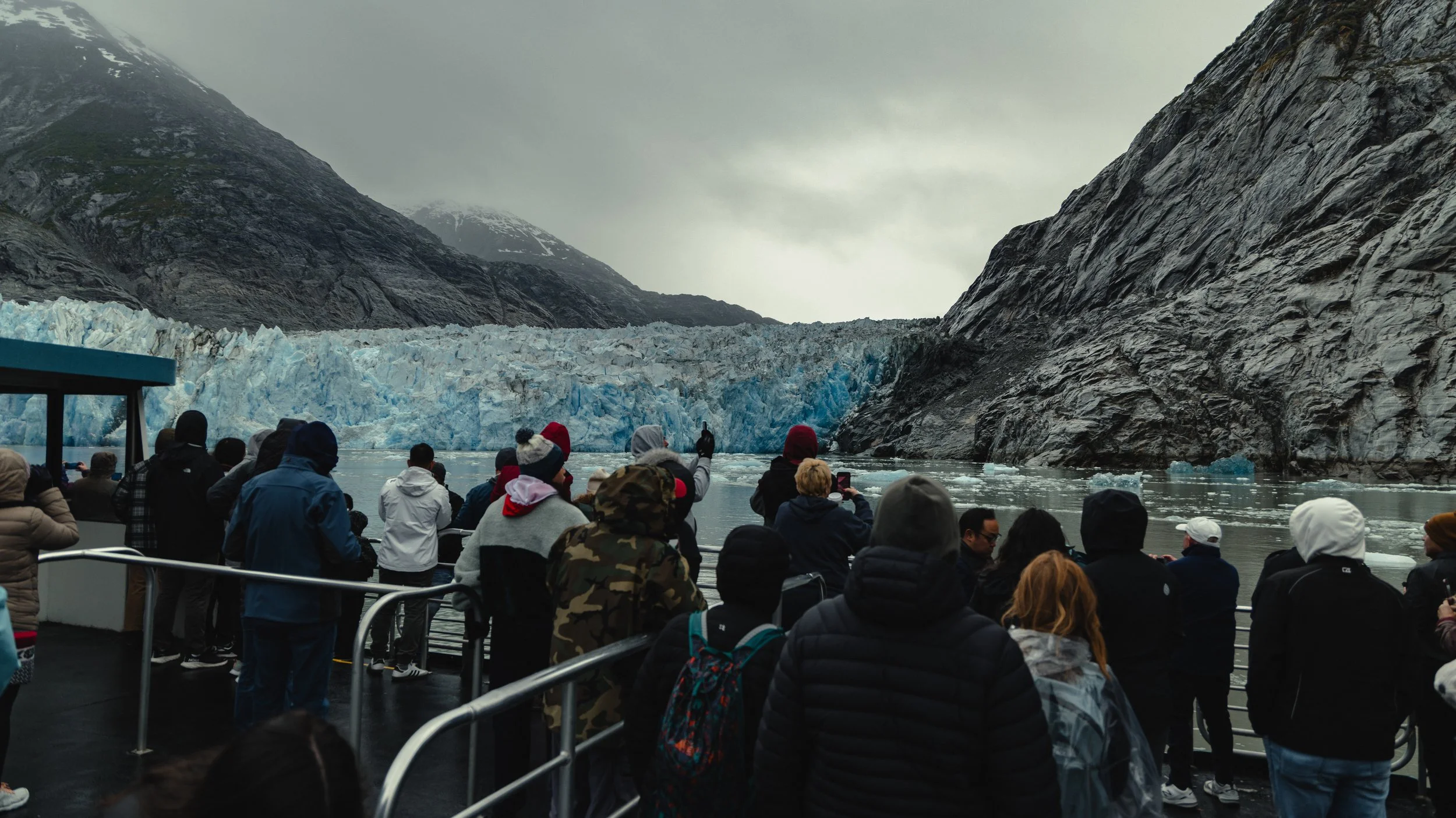 A group of people on a boat viewing a glacier between two mountains on a cloudy day.