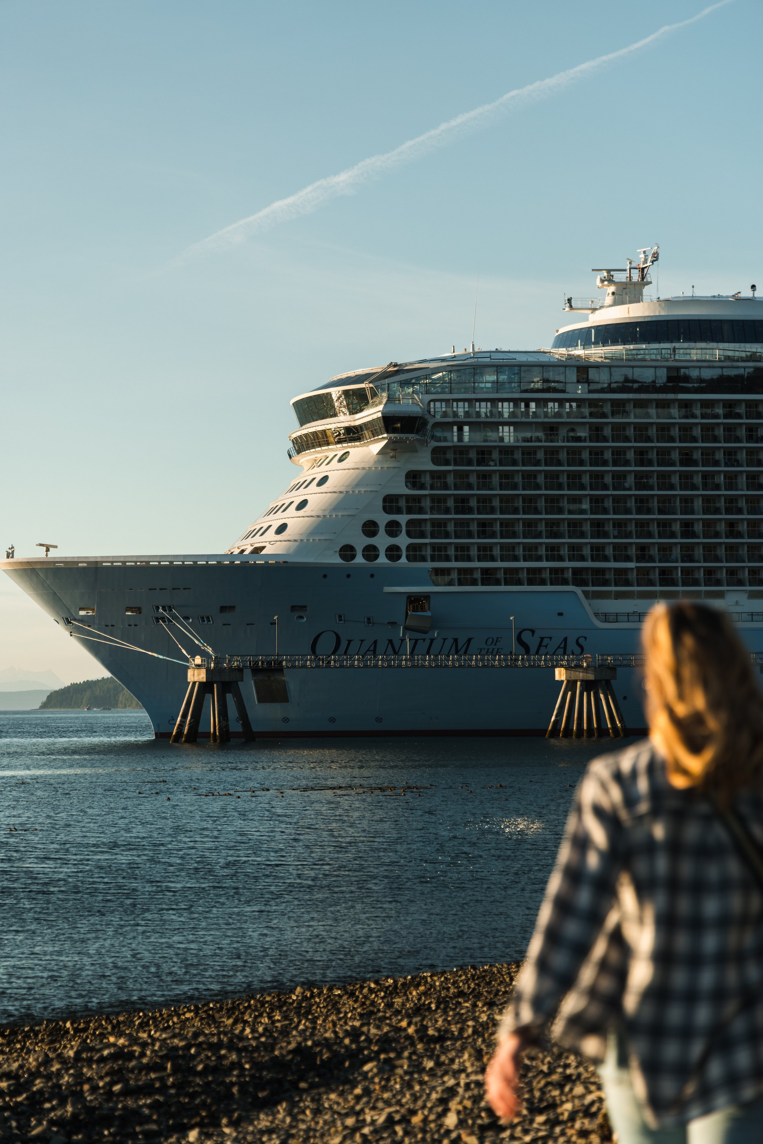A person wearing a plaid shirt sitting by the water's edge with a large cruise ship, Quantum of the Seas, docked nearby at a harbor during sunset.