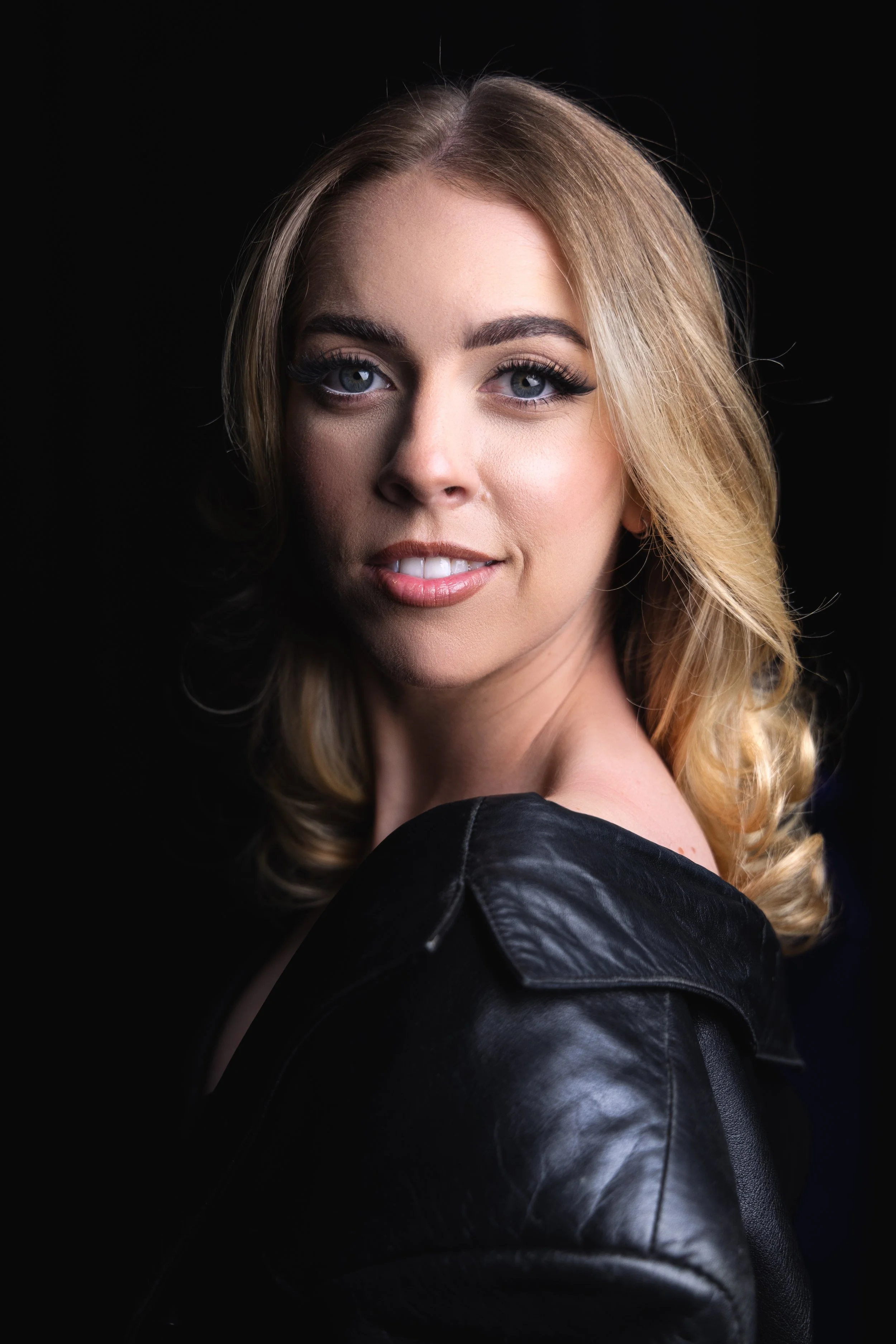 Close-up portrait of a woman with blonde hair, blue eyes, and light makeup wearing a black leather jacket against a black background.