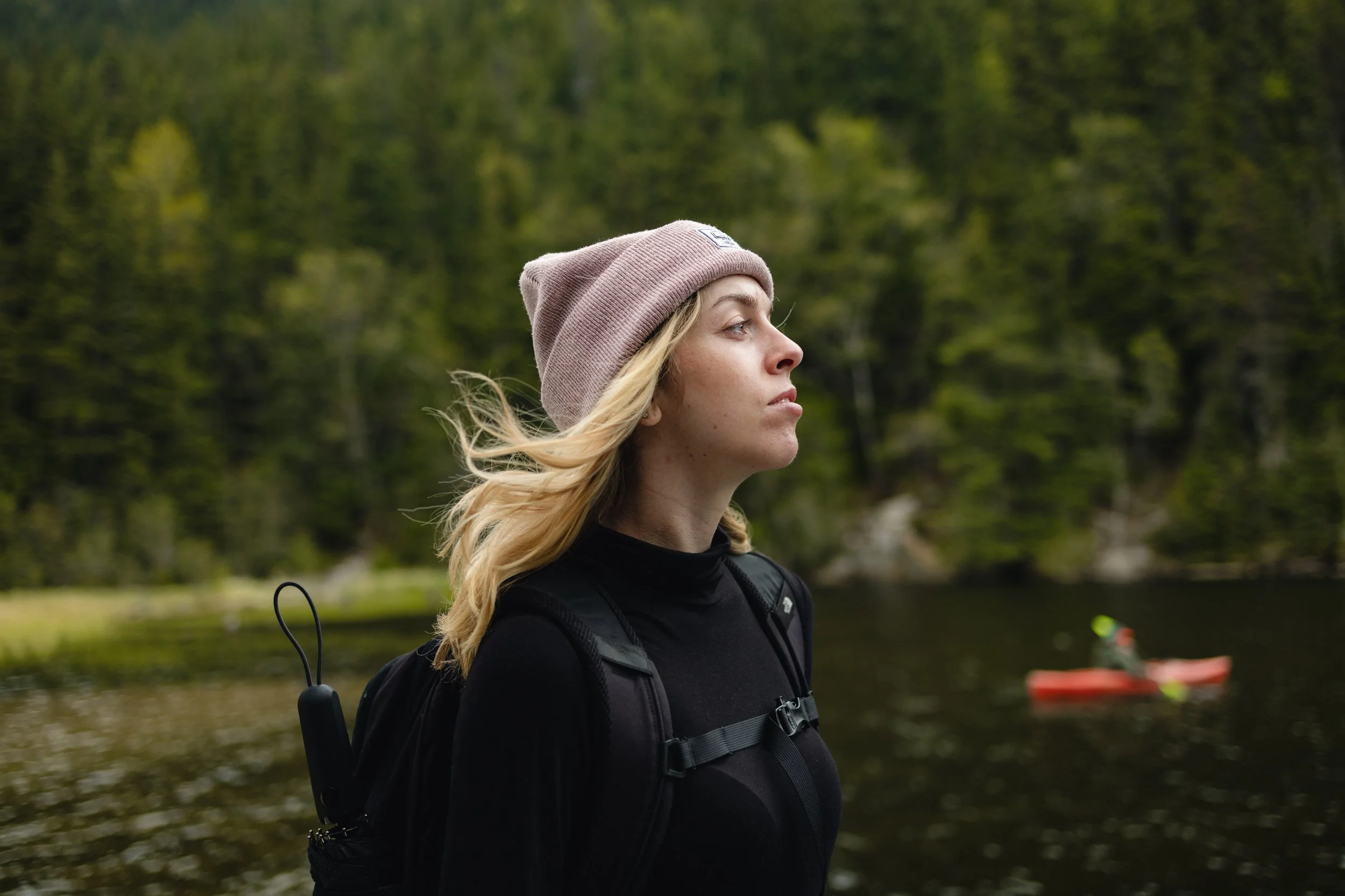 A young woman with blonde hair, wearing a pink beanie and black jacket, standing near a body of water with a forest background.