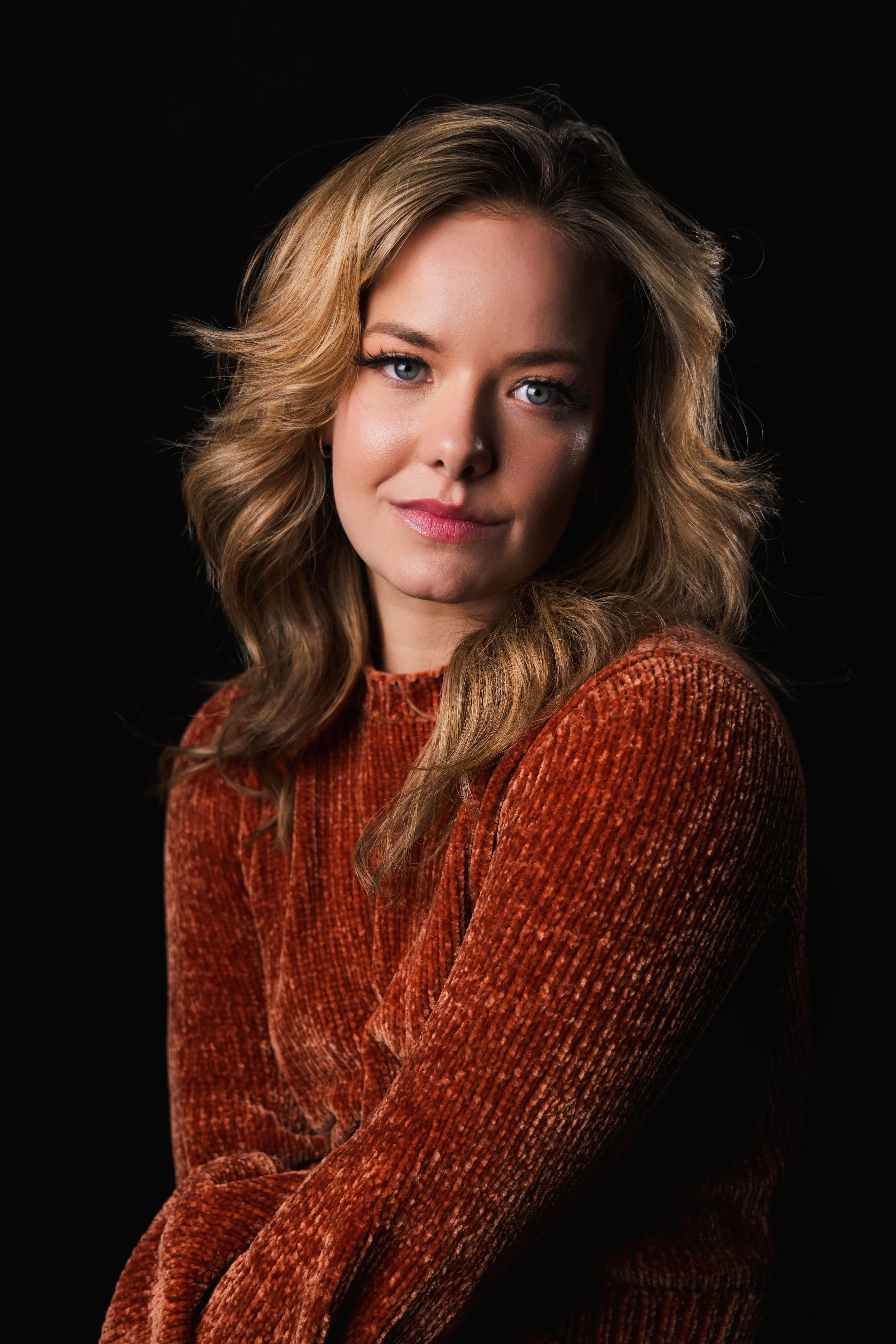 Portrait of a young woman with wavy blonde hair, wearing a rust-colored textured sweater, against a black background.