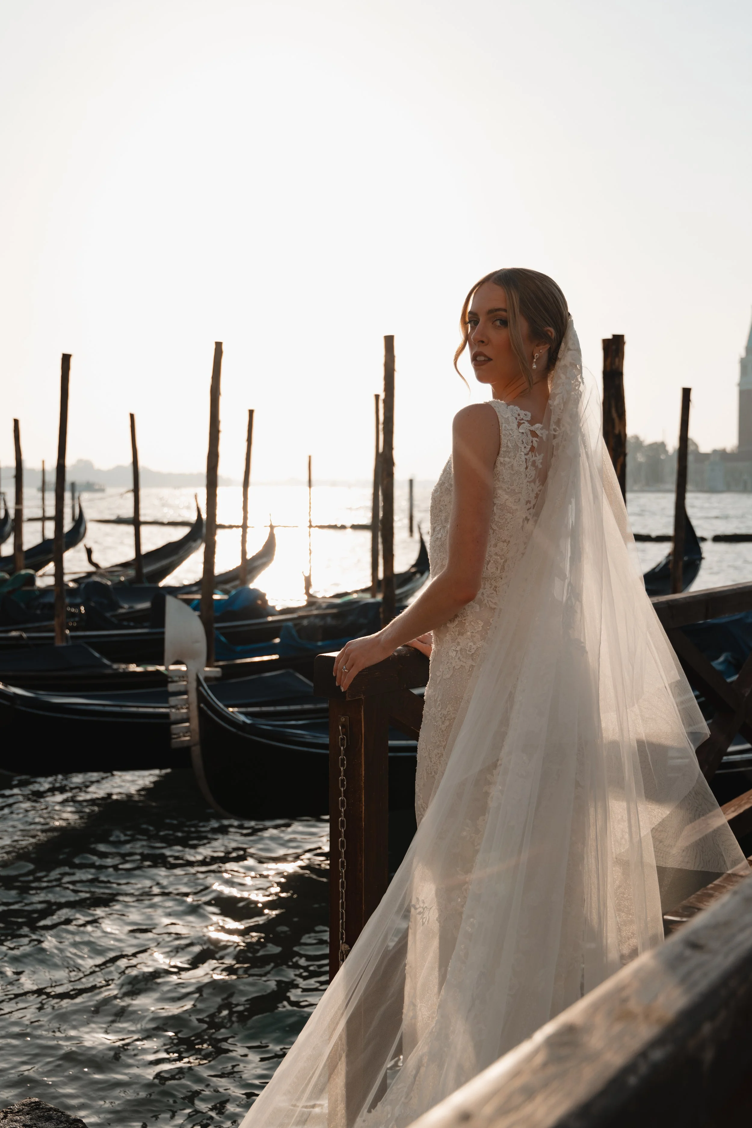 A bride in a lace wedding dress and veil standing on a dock overlooking gondolas on a river, with the setting sun reflected in the water.
