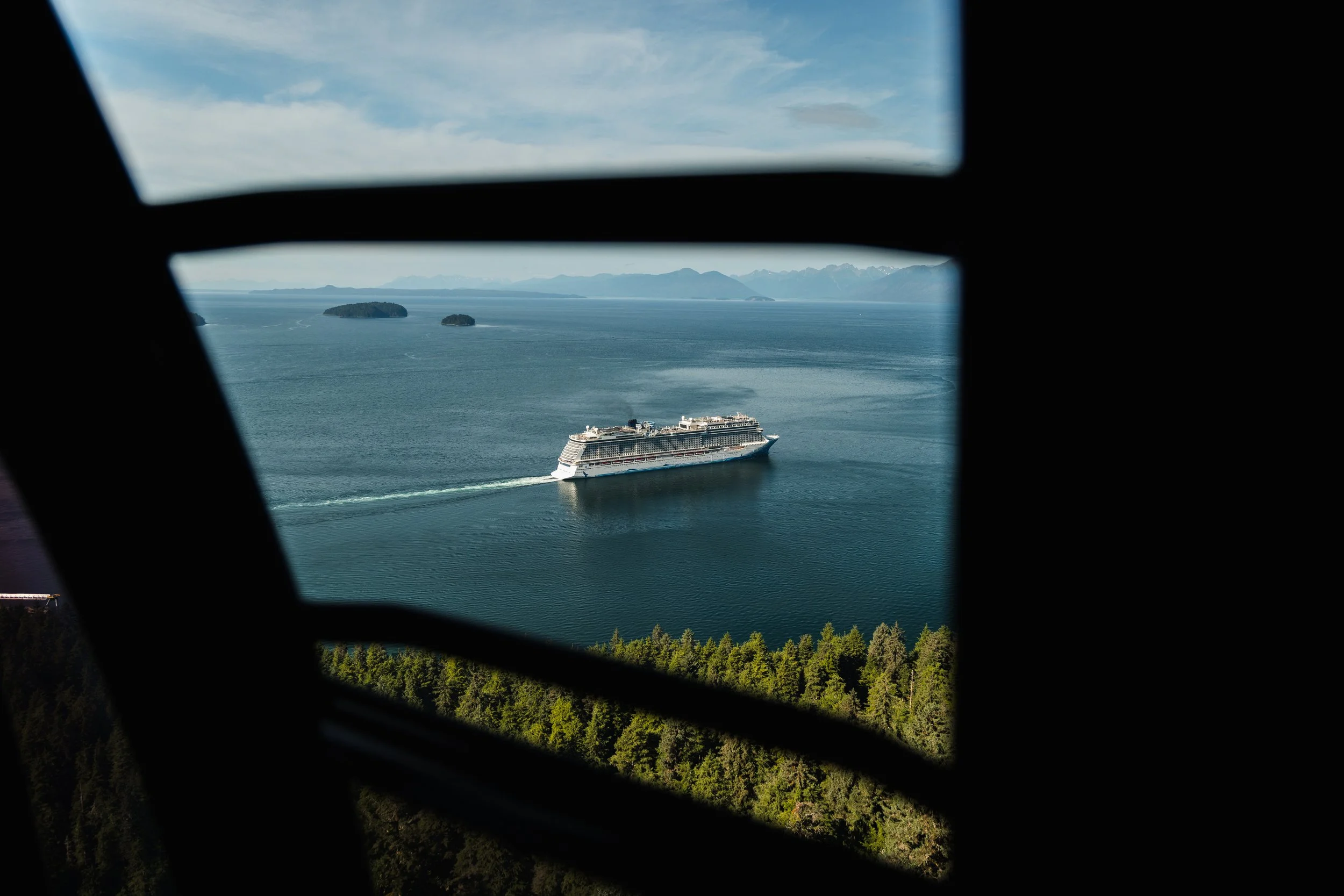 Photo taken from inside a vehicle showing mountains, islands, a cruise ship, water, and a cloudy sky.