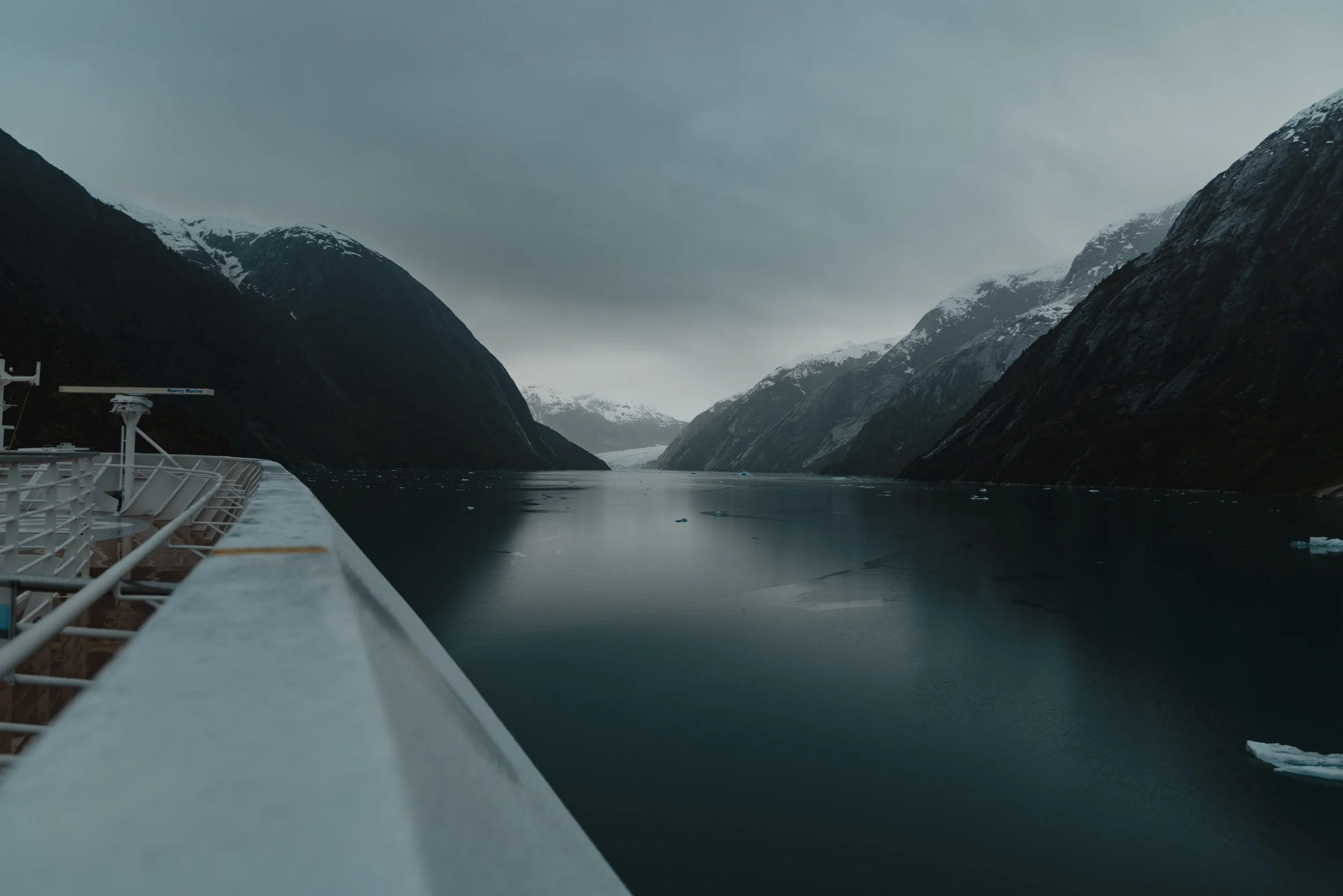 View from a cruise ship sailing through a narrow fjord surrounded by steep, snow-capped mountains on a cloudy day.
