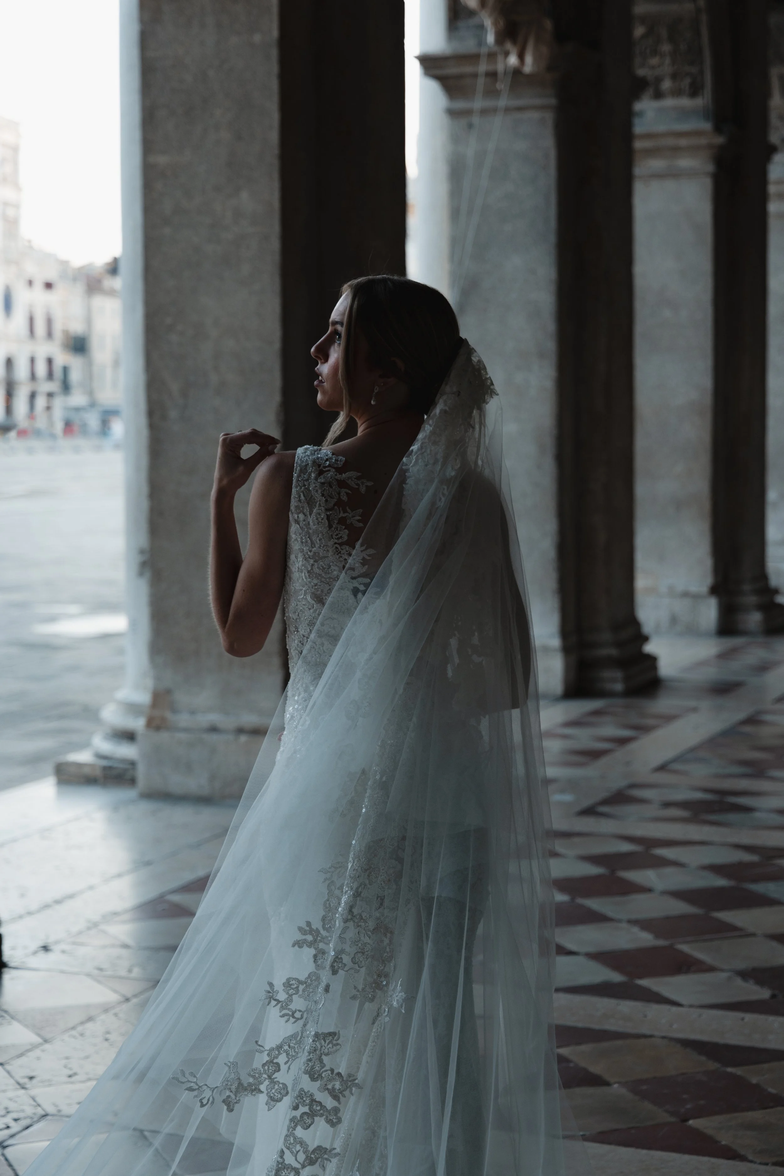 A woman in a wedding dress with lace details is standing inside a building near the columns, looking to the side with her hand near her face. She is wearing a veil and earrings, with a cityscape visible through the open doorway behind her.