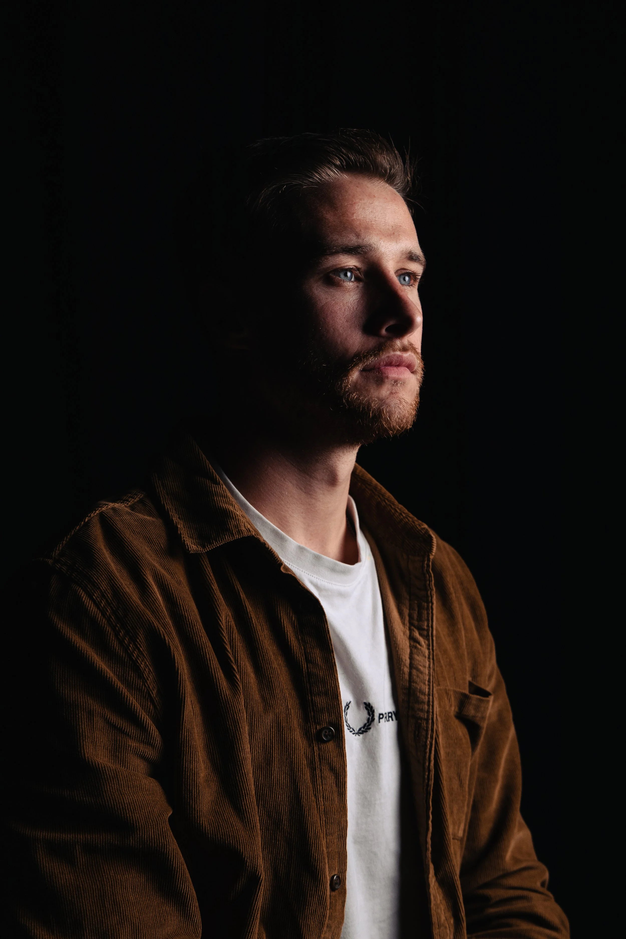 A young man with blonde hair and a beard, wearing a brown corduroy jacket over a white T-shirt, looking to his right against a black background.