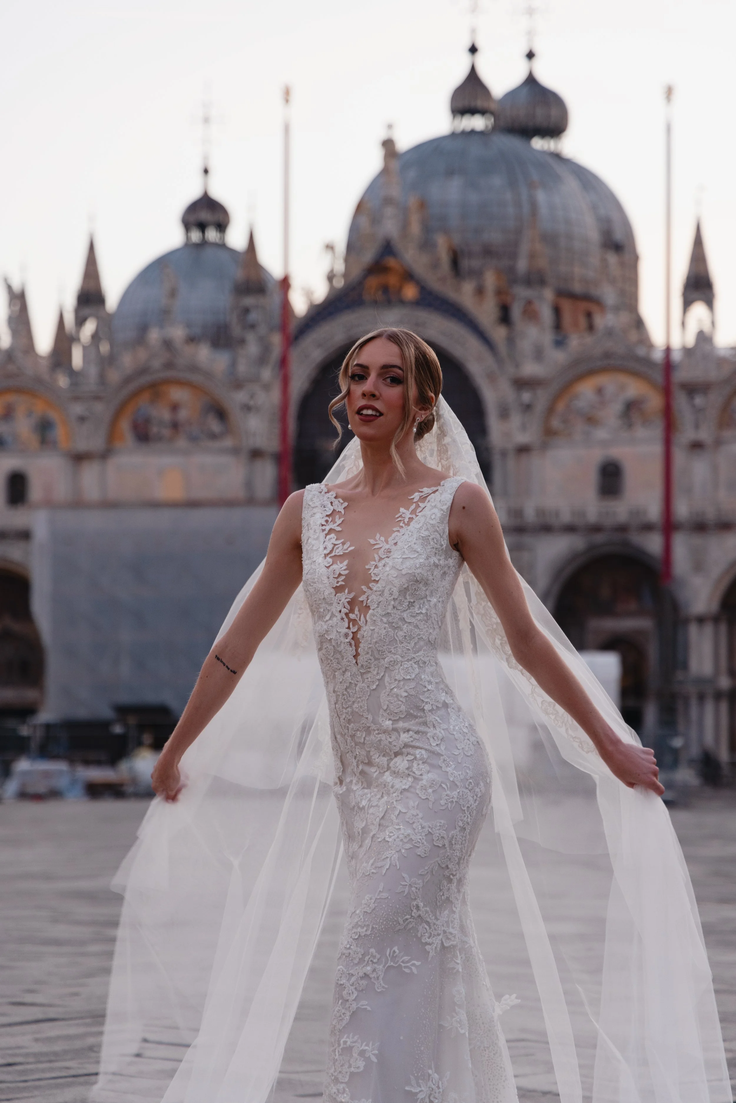 A bride in a lace wedding gown with a tulle train standing outdoors in front of historic buildings with domed roofs, possibly in Venice, Italy.