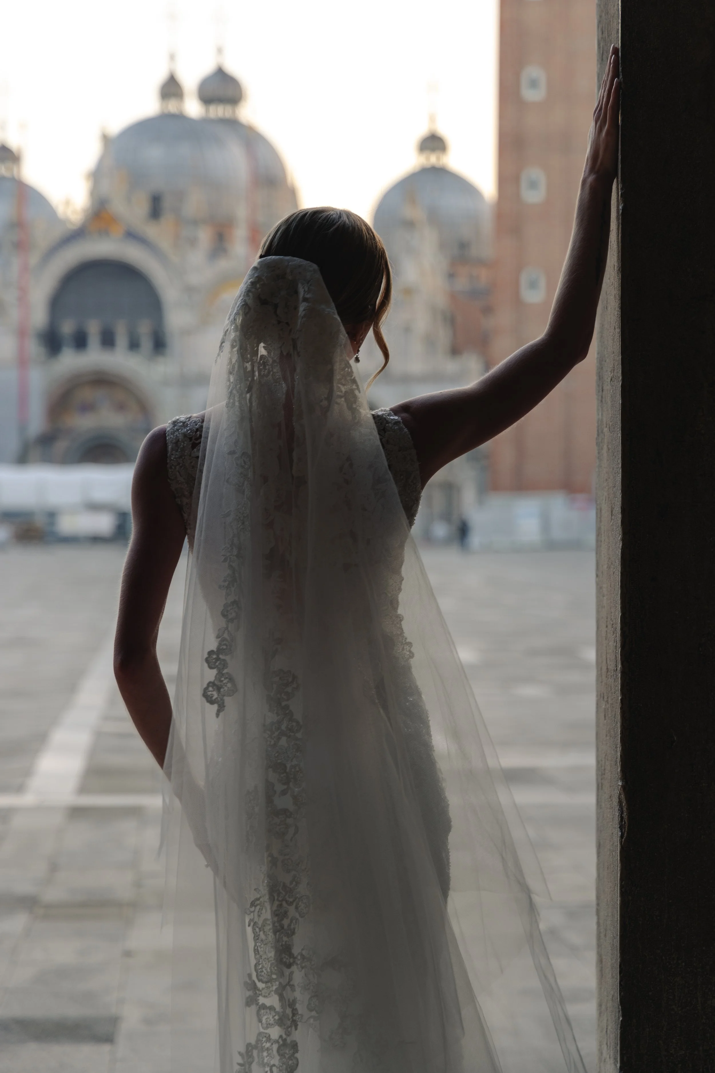 A woman in a wedding dress and veil is standing near a wall with her hand resting on the wall. She is looking away from the camera, with her back to the viewer. In the background, there is an ornate building with domed rooftops and spires.