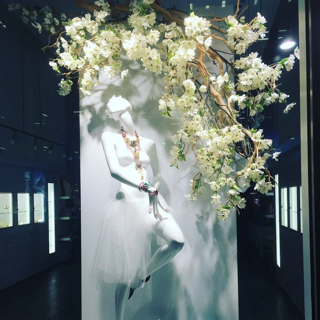 A mannequin adorned with colorful jewelry is positioned in a jewelry store display, surrounded by white flowers and branches hanging above.
