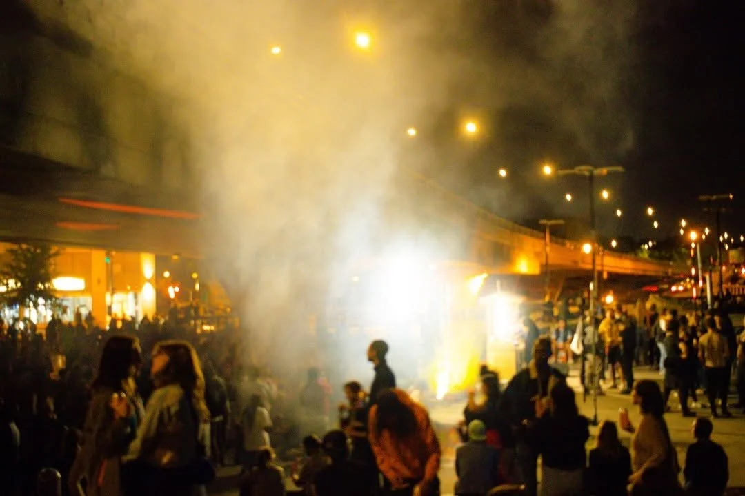Crowd of people gathered at night with bright lights, smoke, and some street vendors or stalls.