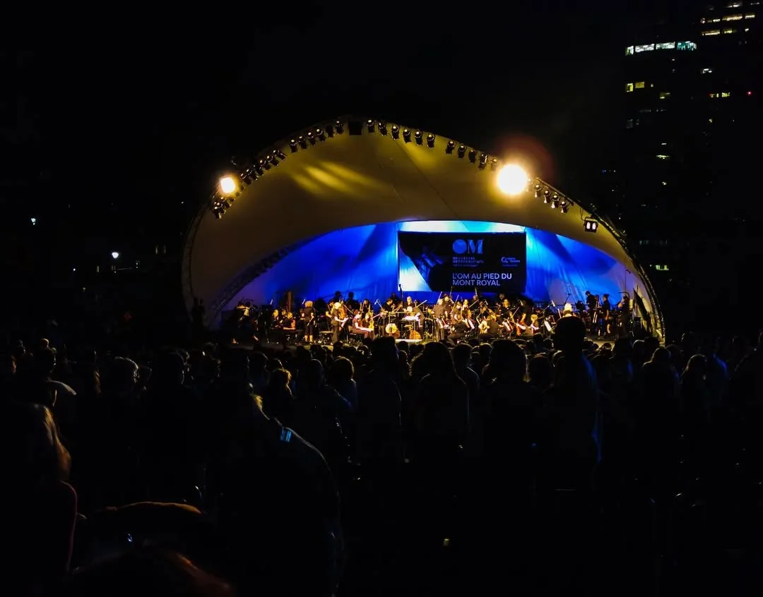 An outdoor concert at night with a large crowd watching an orchestra perform on a stage with a curved canopy and blue lighting.